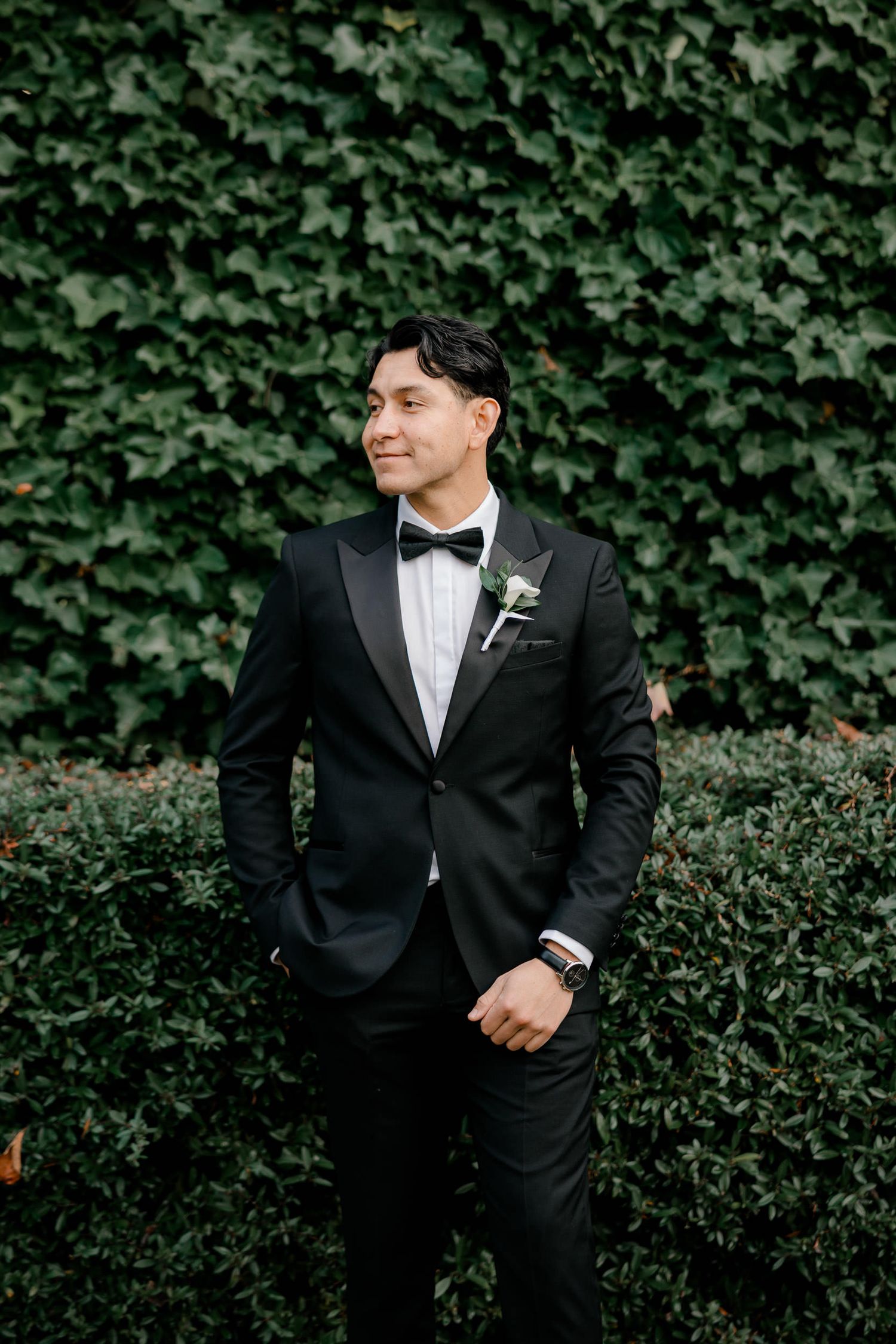 A groom in a classic black tuxedo and bow tie poses against a lush green hedge backdrop.