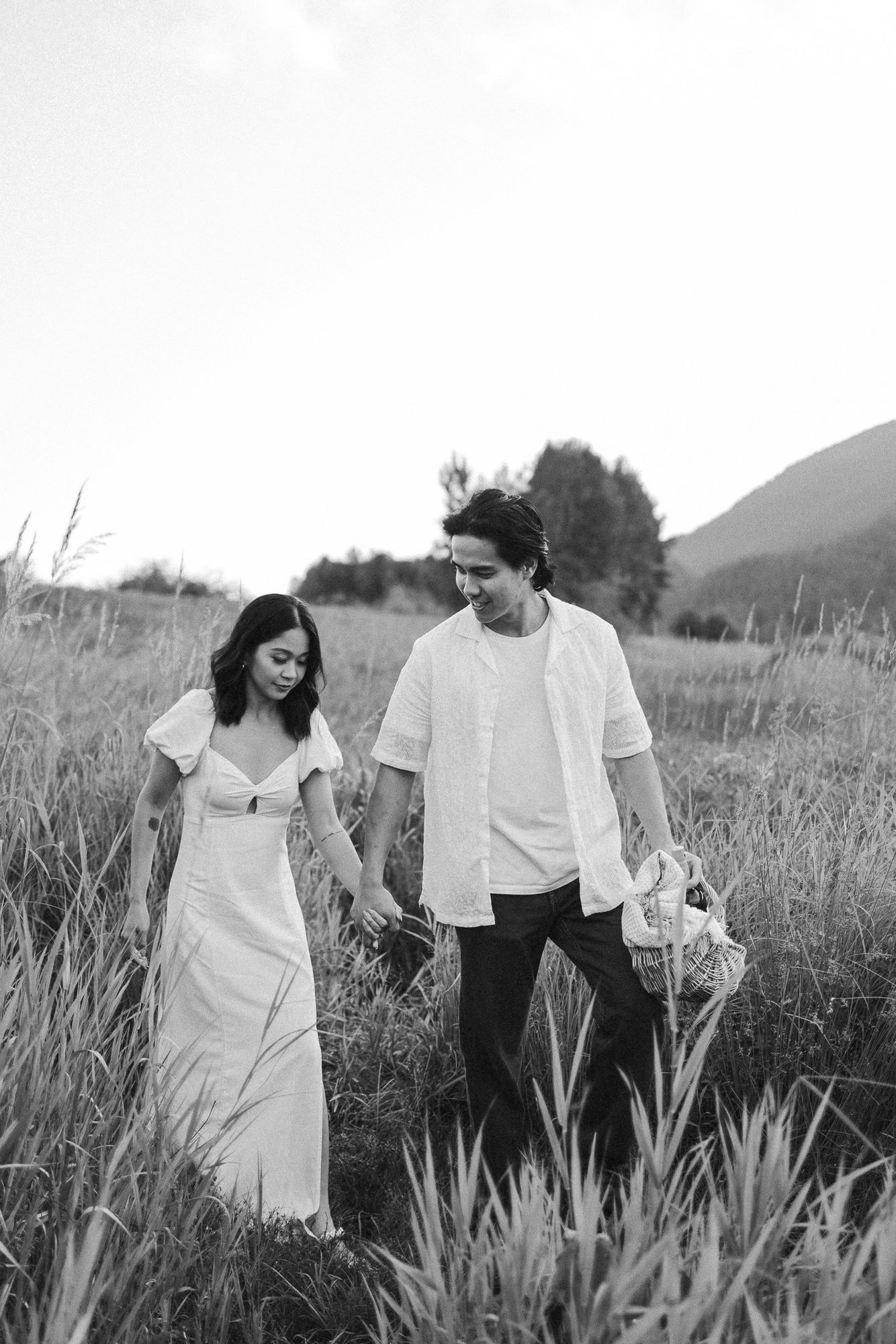 Black and white photo of a couple holding hands and walking through tall grass with a picnic basket near mountains.