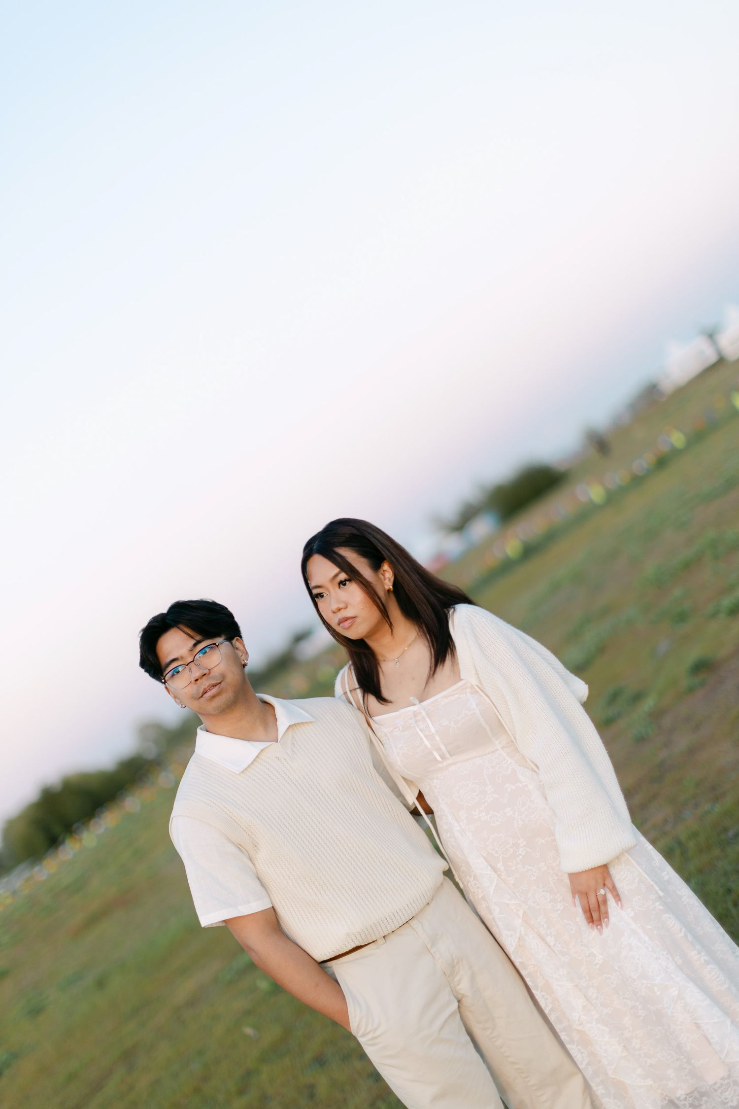 Couple in cream outfits standing close together in a grassy field at sunset during an engagement photo session.