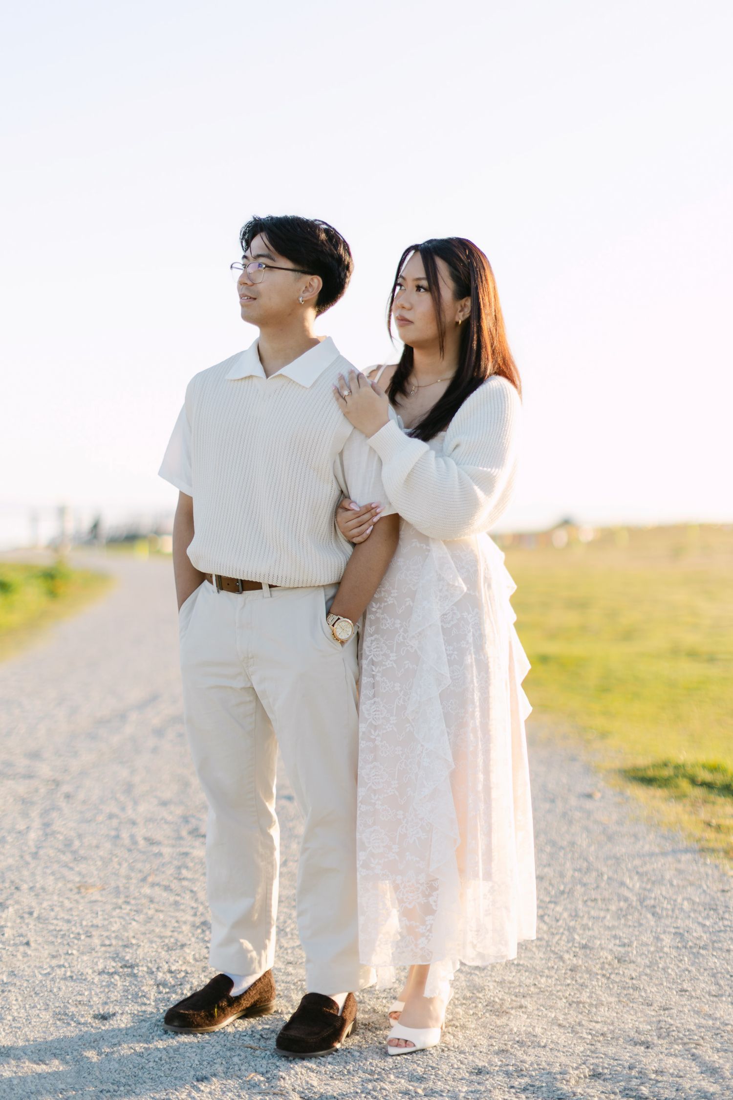 Couple in matching white outfits posing on a gravel path at sunset, her arm wrapped around his as they gaze into the distance.