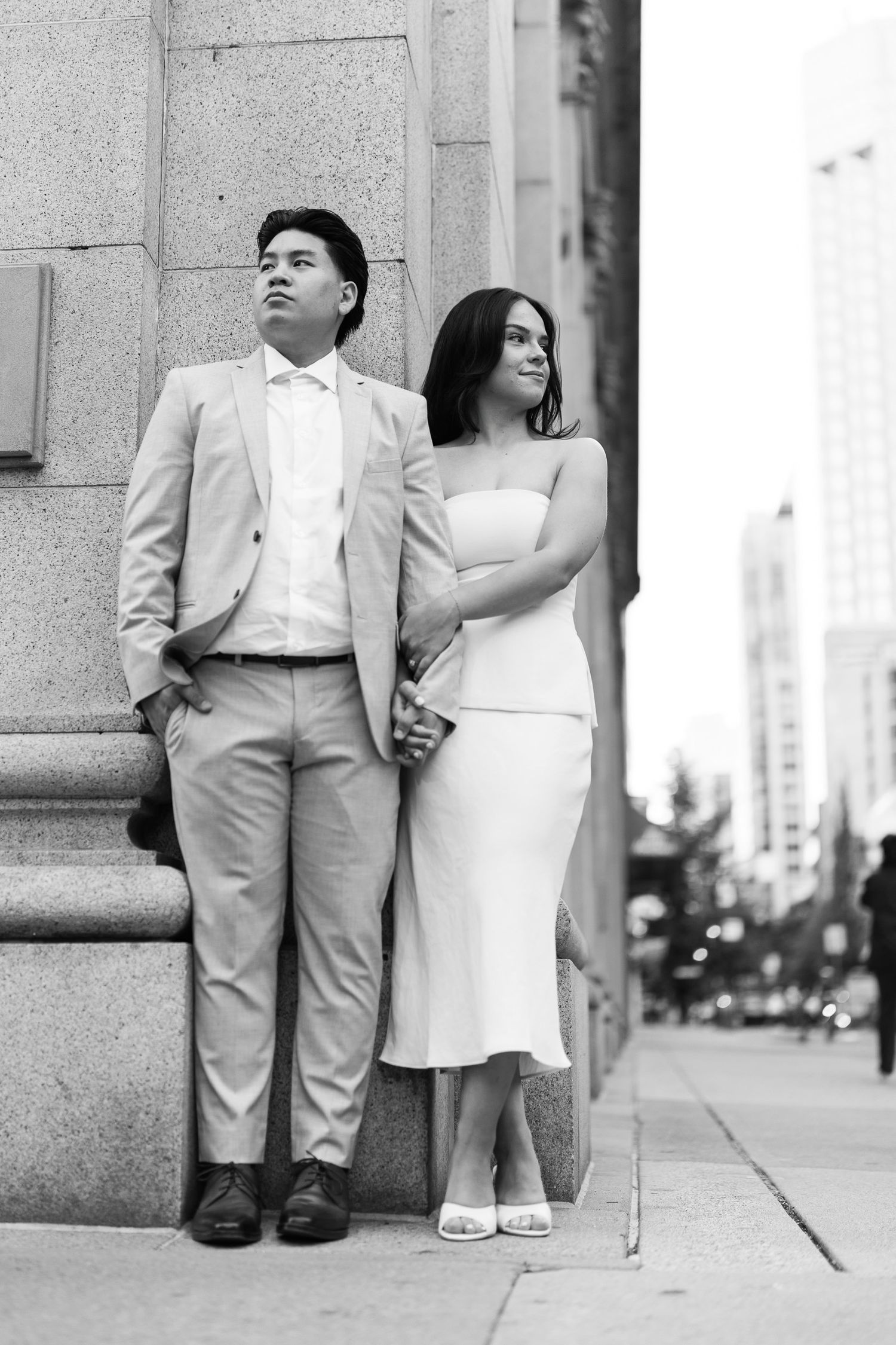 Black and white photo of a couple holding hands and looking away as they lean against a stone building on a city sidewalk.