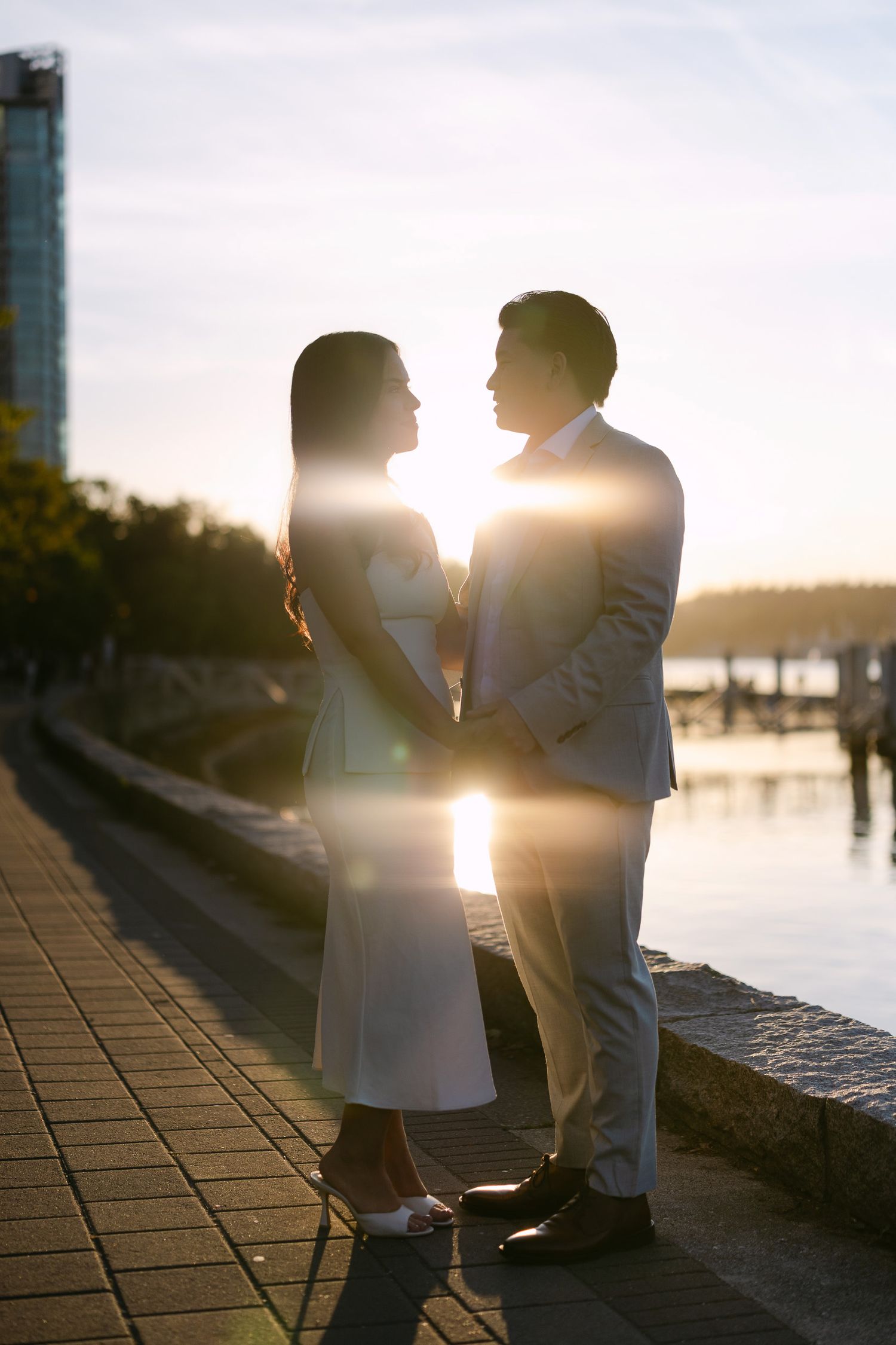 Couple in elegant attire share a romantic moment facing each other on a waterfront promenade at golden hour with sun flare.