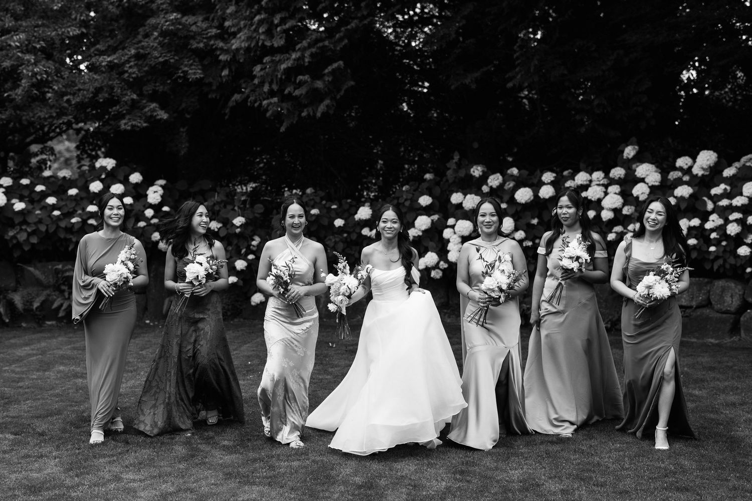 Bride and bridesmaids in elegant gowns pose together outdoors with hydrangeas in black and white wedding photo.
