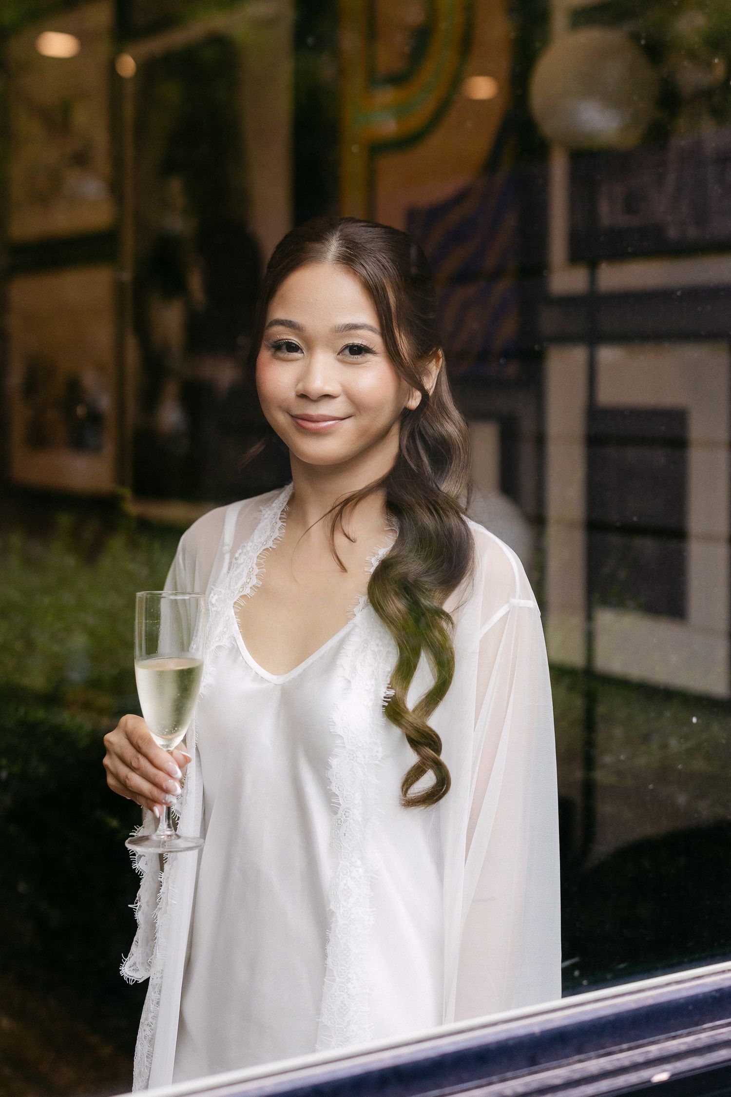 A young woman in a white satin robe holds a champagne flute, smiling softly with wavy hair.