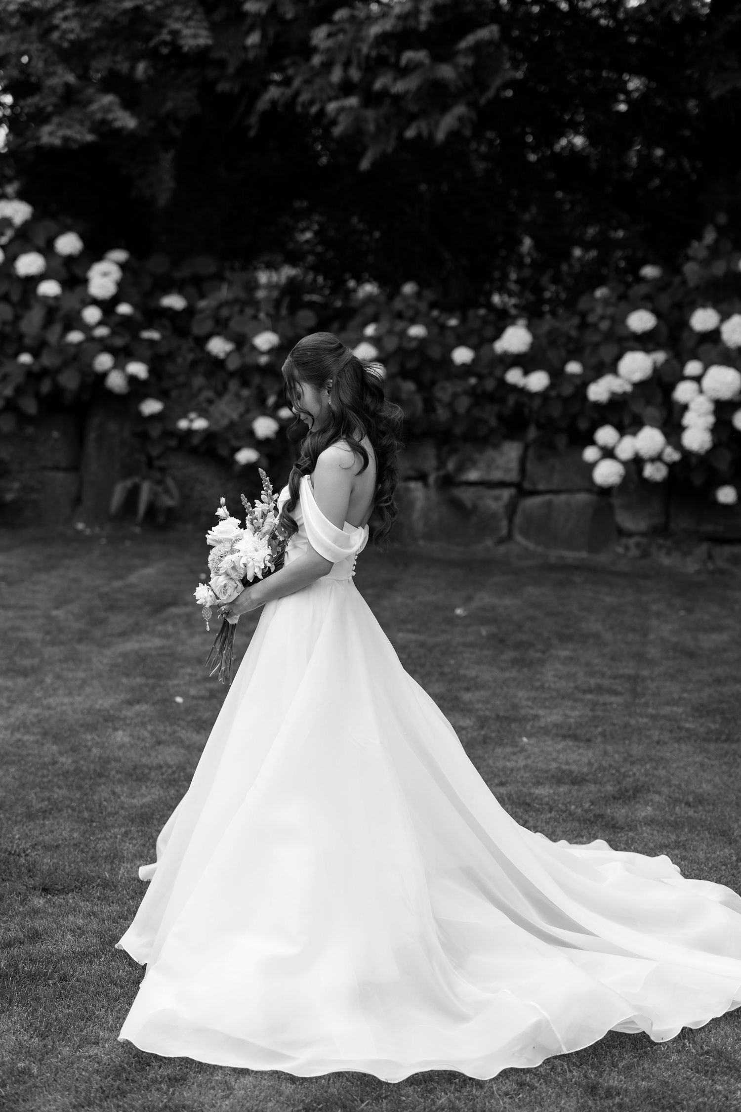 Black and white photo of a bride in an off-shoulder ball gown holding a bouquet in a garden.