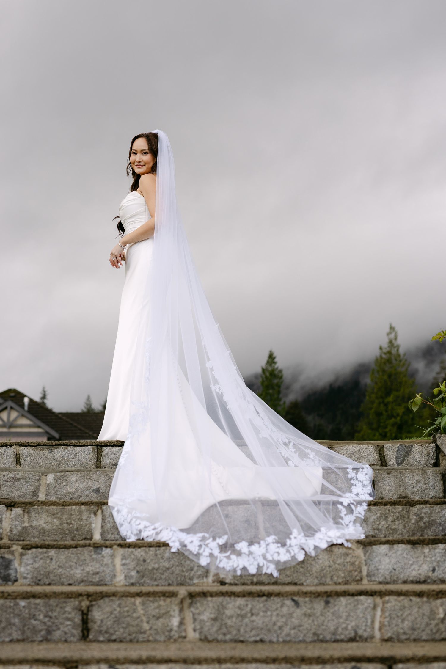 Bride in white gown and long lace-trimmed veil stands on stone steps with misty mountain backdrop.