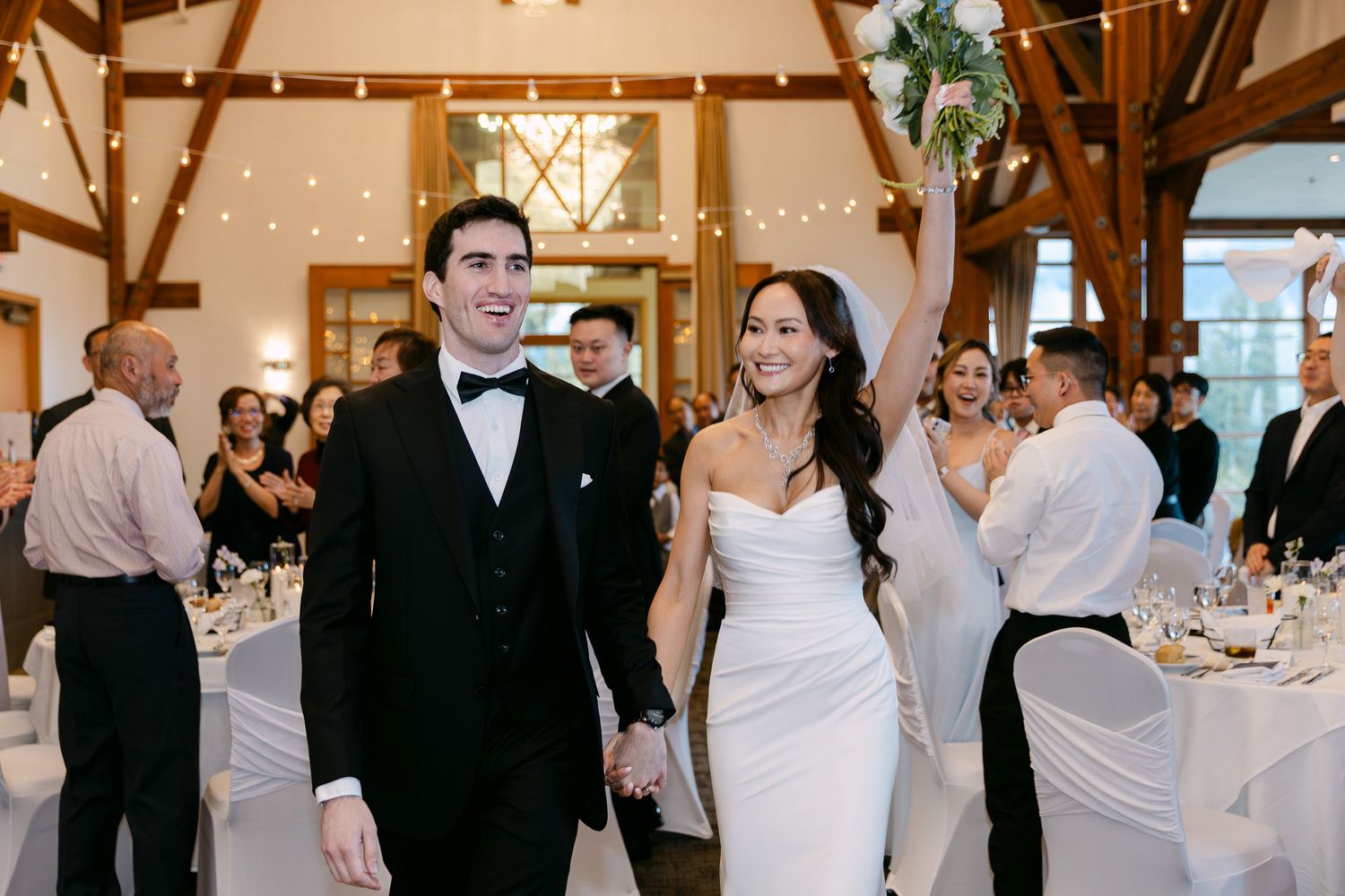 Bride and groom entering wedding reception hall, holding hands and smiling, guests applauding in elegant venue with string lights.