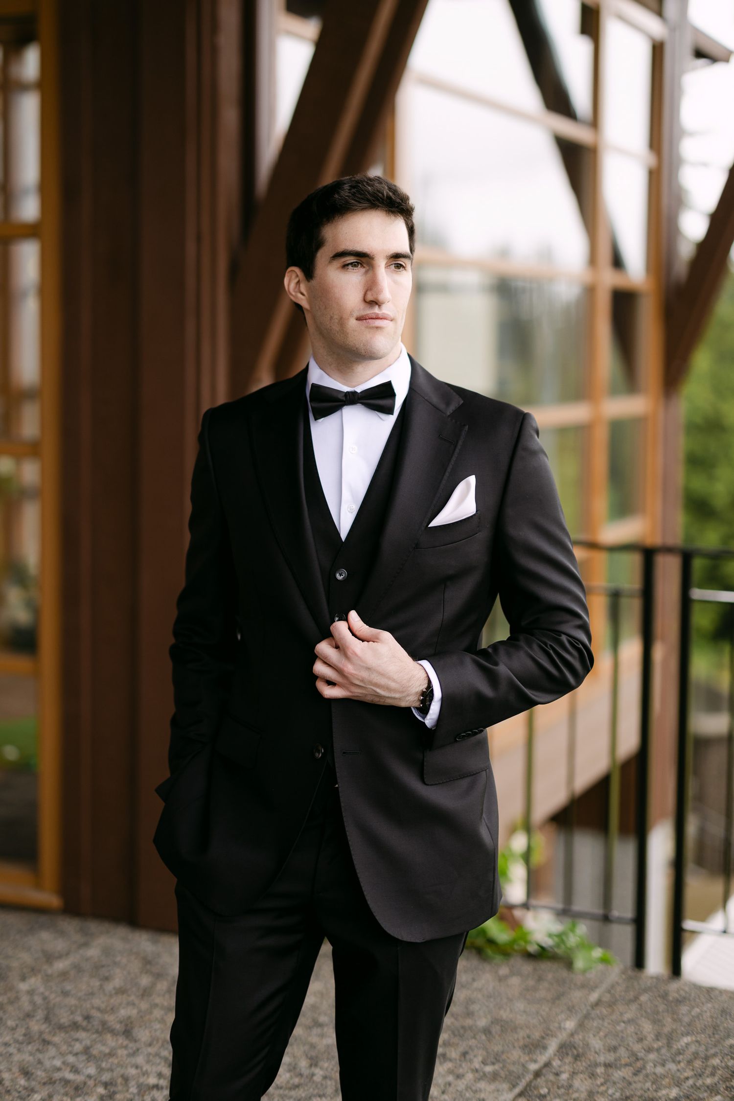 A dapper groom poses in a classic black tuxedo with bow tie and white pocket square outside a modern venue.