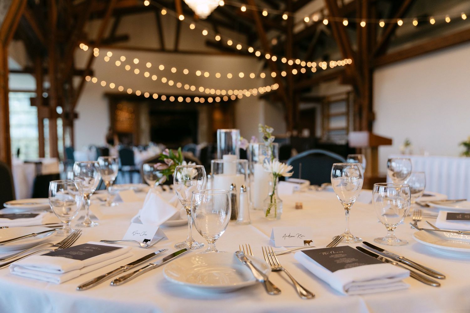 Elegant wedding reception table set with white linens, glassware, and string lights overhead.
