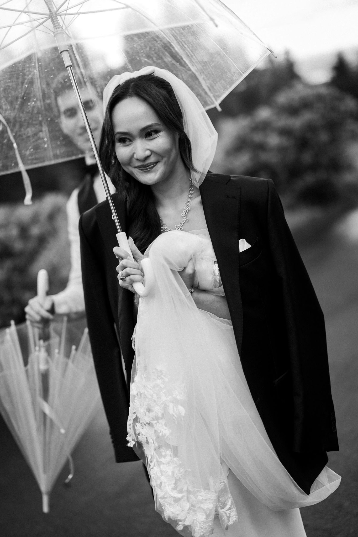 Black and white photo of smiling bride in veil and grooms jacket holding clear umbrella in the rain.