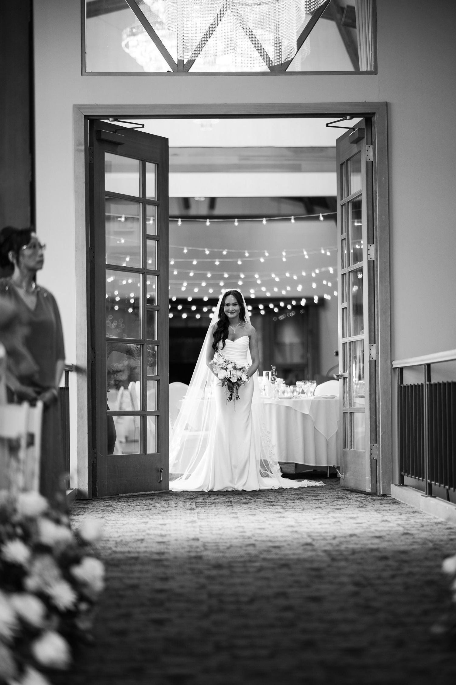 Bride in white gown stands in French doorway with fairy lights and reception tables behind her.
