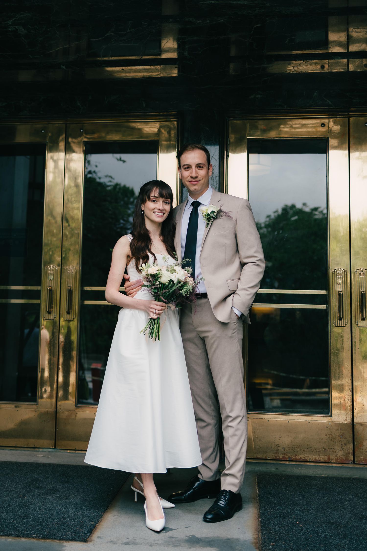 A bride in a white dress and groom in tan suit stand together outside modern building doors.