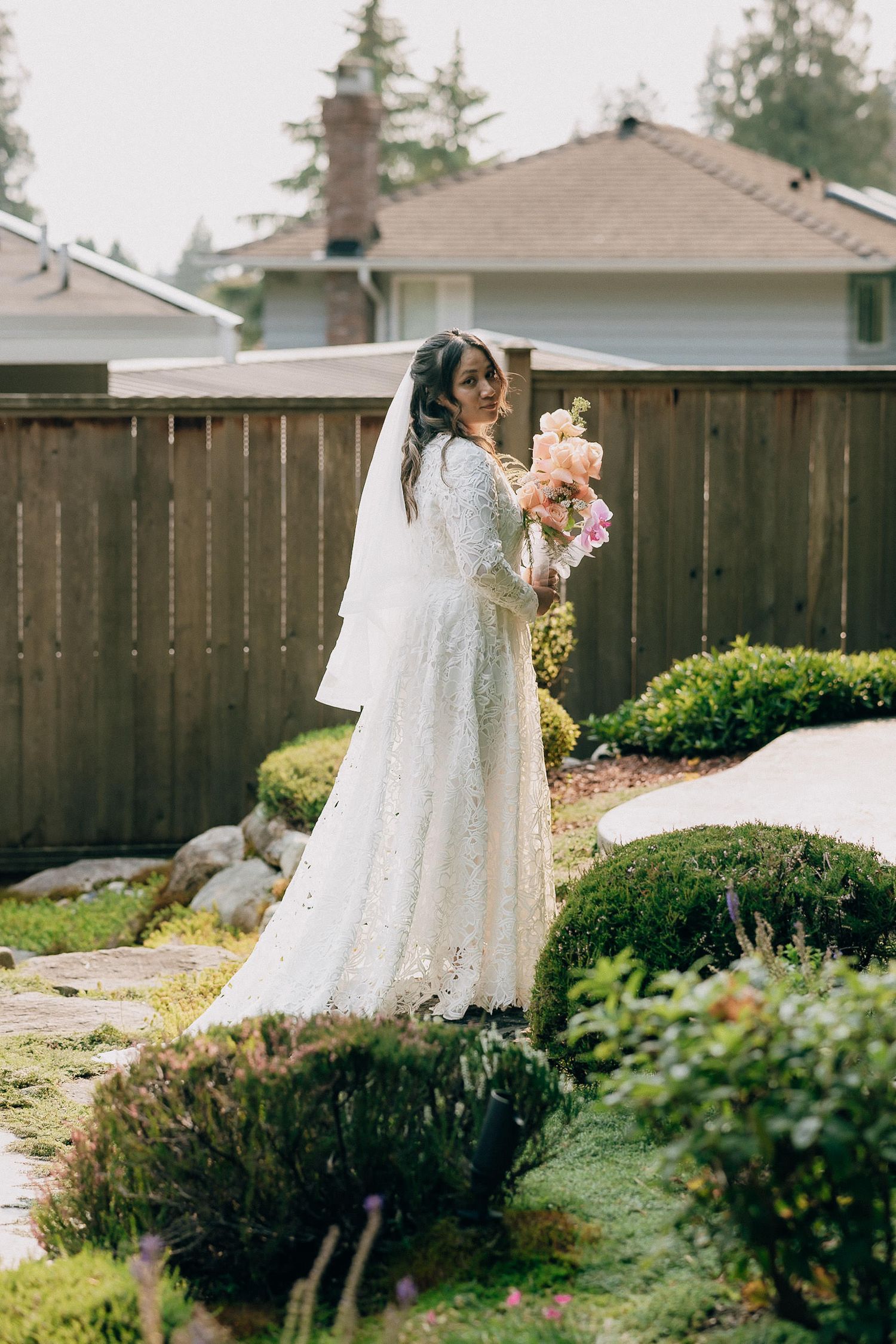 A bride in a flowing white lace dress poses in a backyard garden with wooden fencing and greenery.