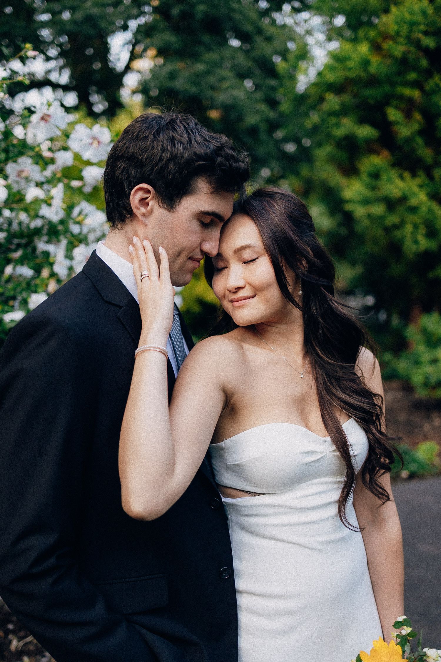 A romantic couple in wedding attire share an intimate moment in a garden setting with lush greenery in the background.