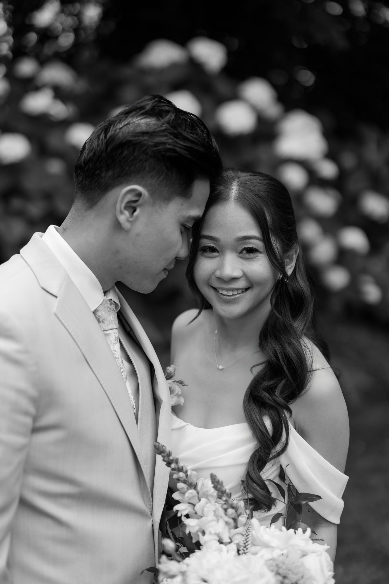 Black and white wedding photo of a smiling bride in white dress with groom in light suit, posing among blooming flowers.