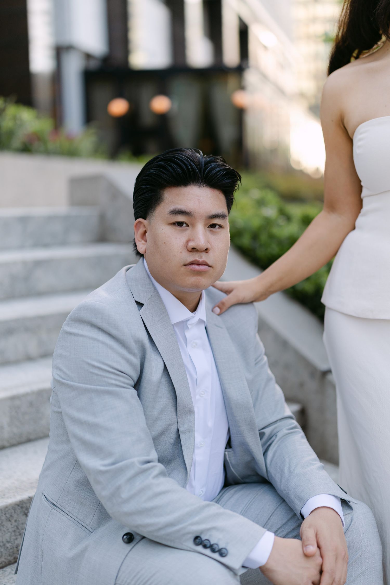 Man in a light gray suit sits on stone steps with a woman in a white dress resting her hand on his shoulder.