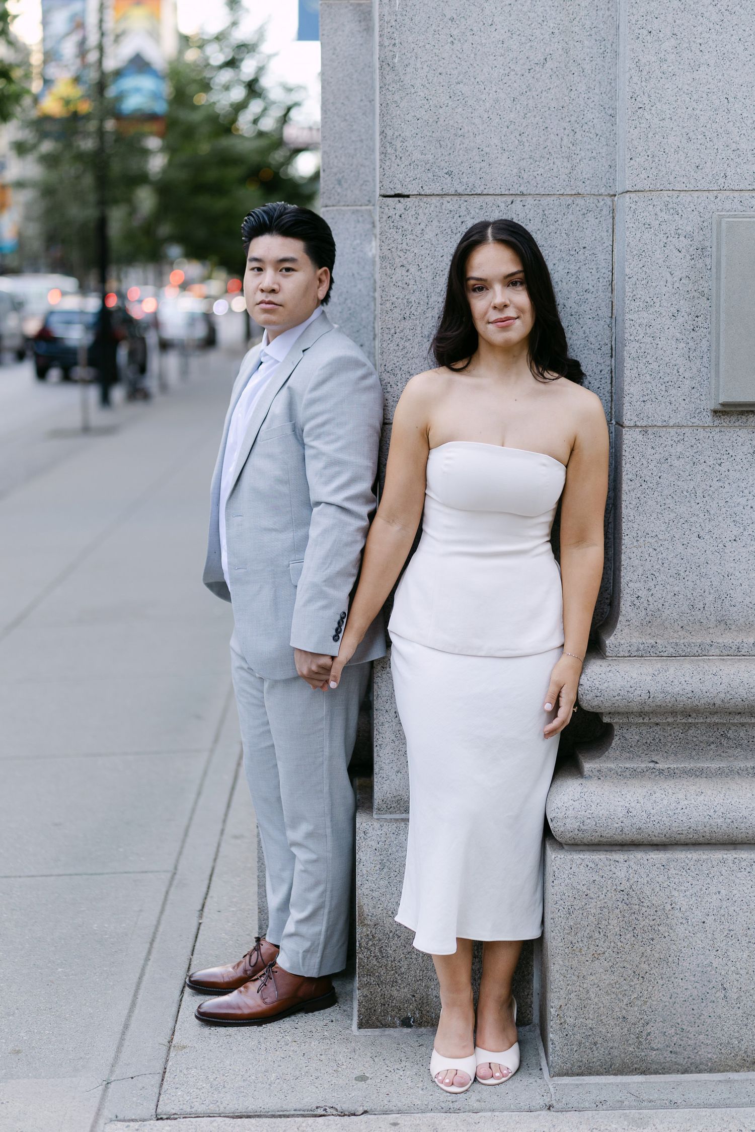 Couple in a gray suit and white dress lean against a granite building column holding hands on a city sidewalk.