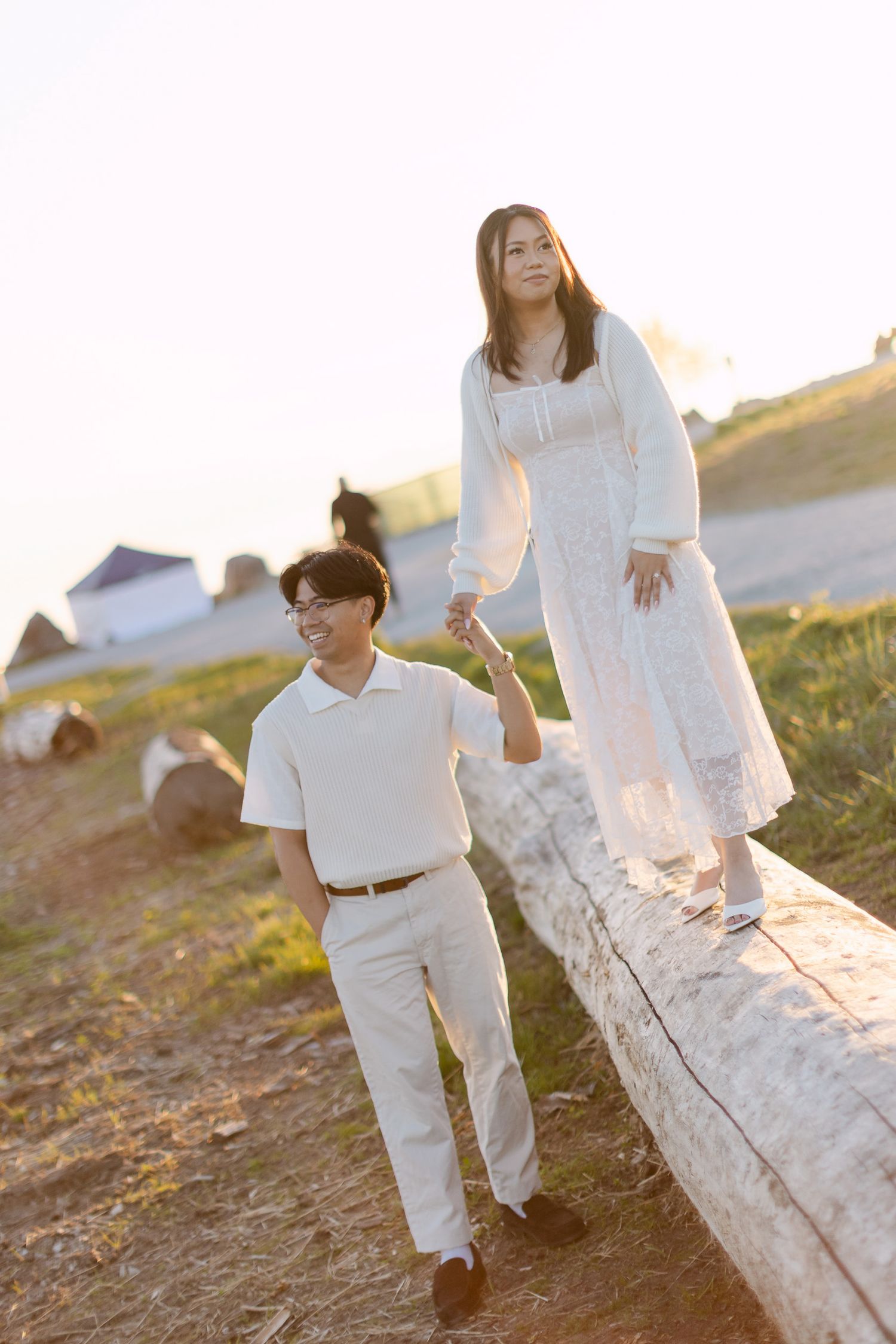 Woman in a white lace dress walks along a driftwood log holding hands with a smiling man in a cream outfit.