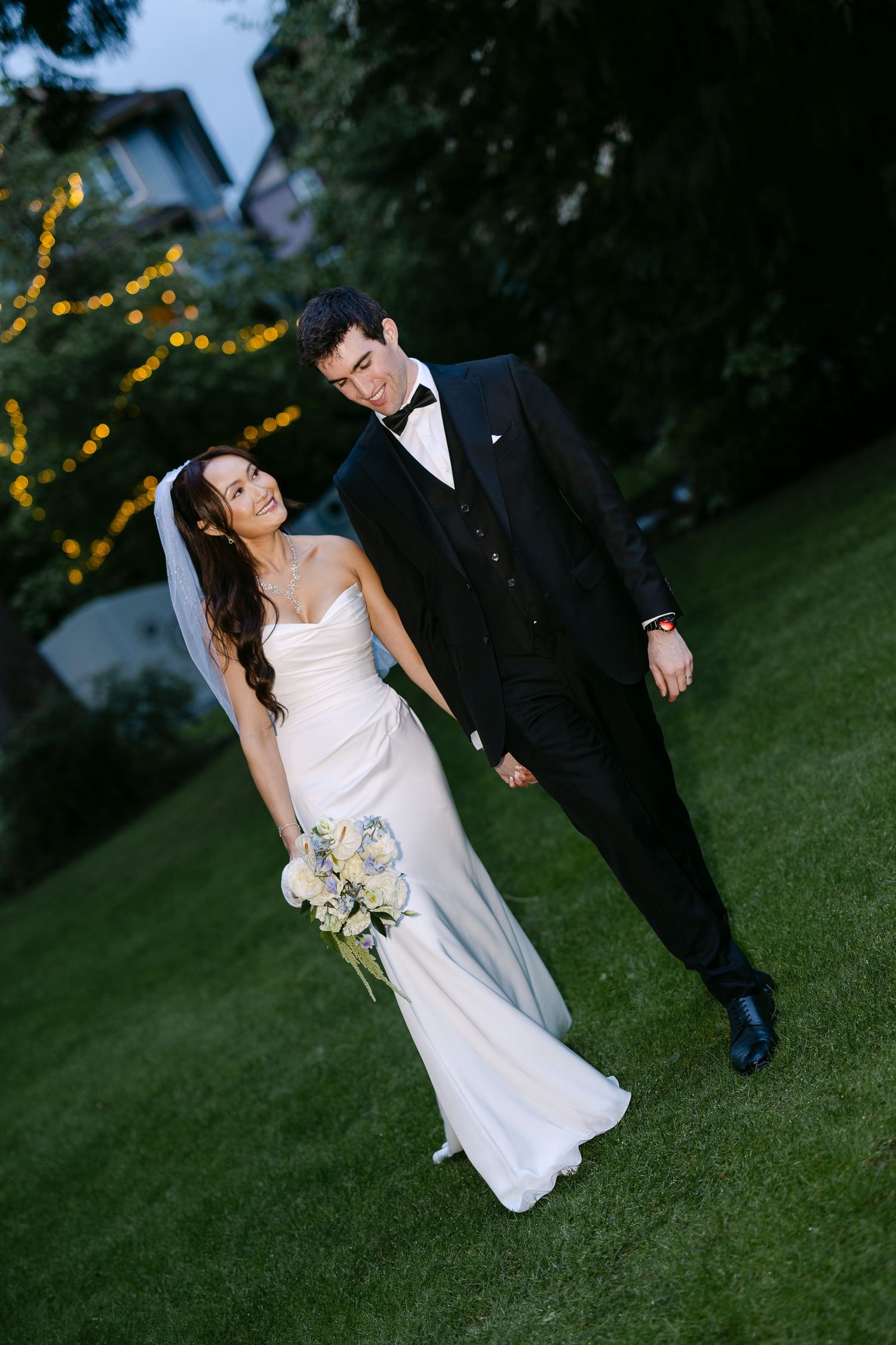 Bride and groom walk hand in hand on green lawn with twinkling lights at dusk.