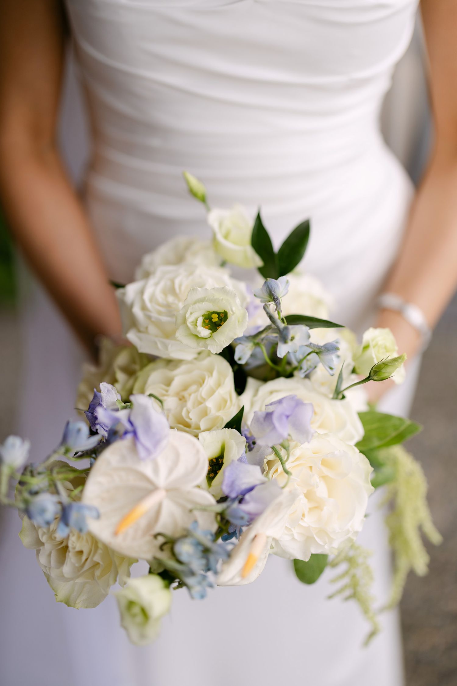 Bride holding a white and lavender wedding bouquet with roses, anthurium, and delicate blue flowers against her white gown.