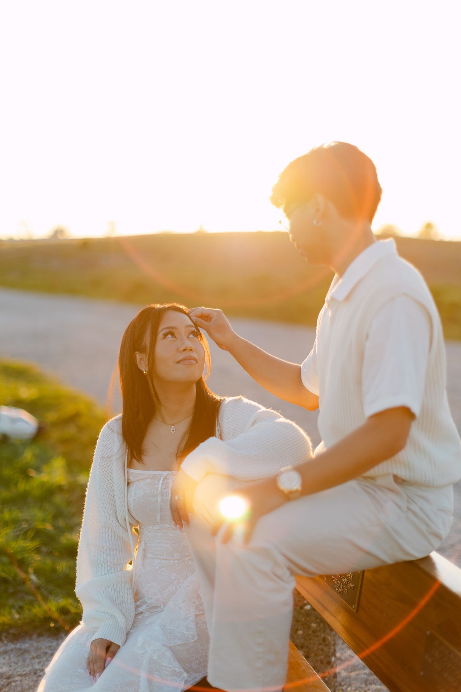 A man in white sits on a bench gently touching a woman's forehead as she looks up at him, bathed in warm golden sunset light outdoors.
