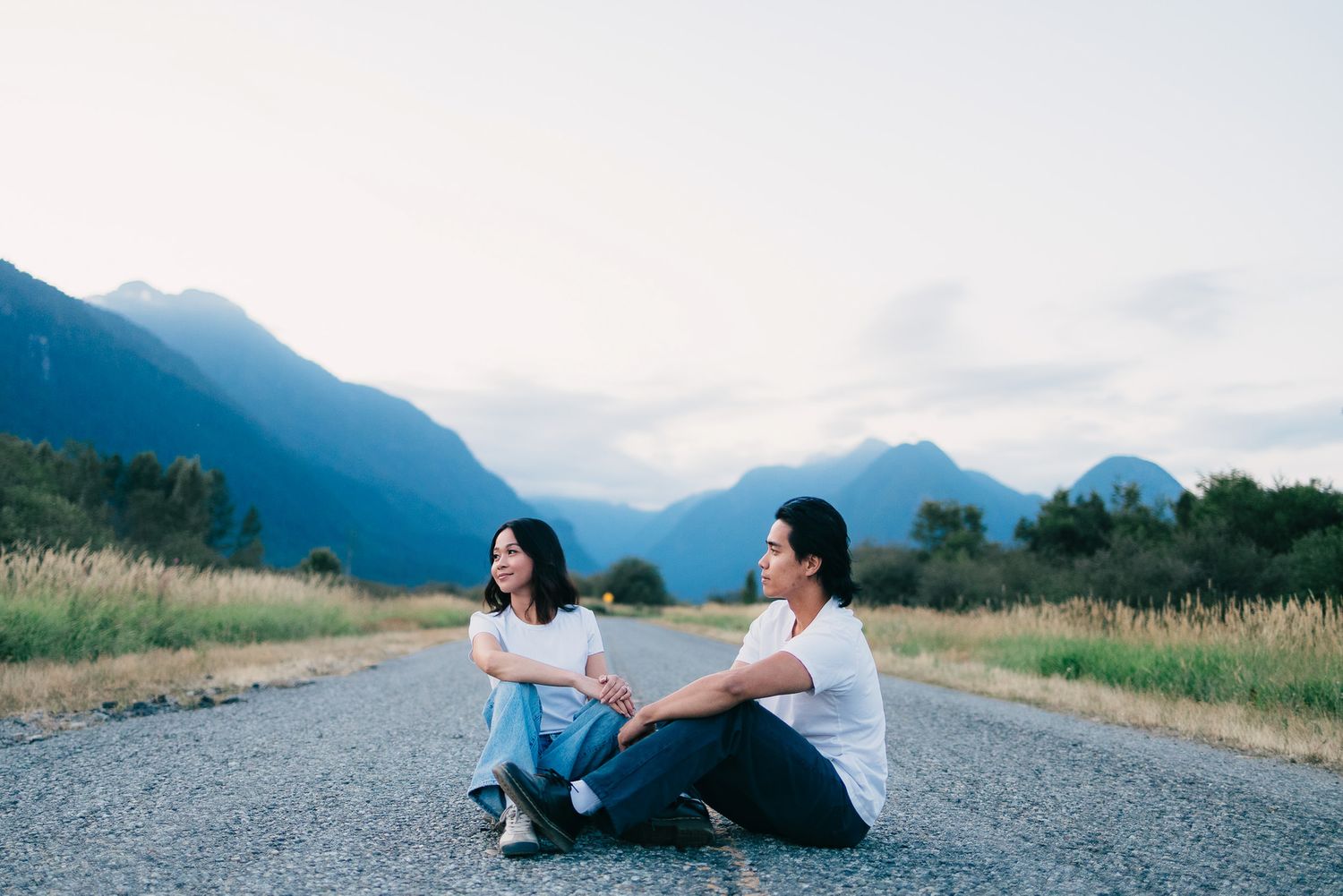 Couple sitting together on an empty paved road holding hands with misty mountains and golden grassy fields in the background at dusk.