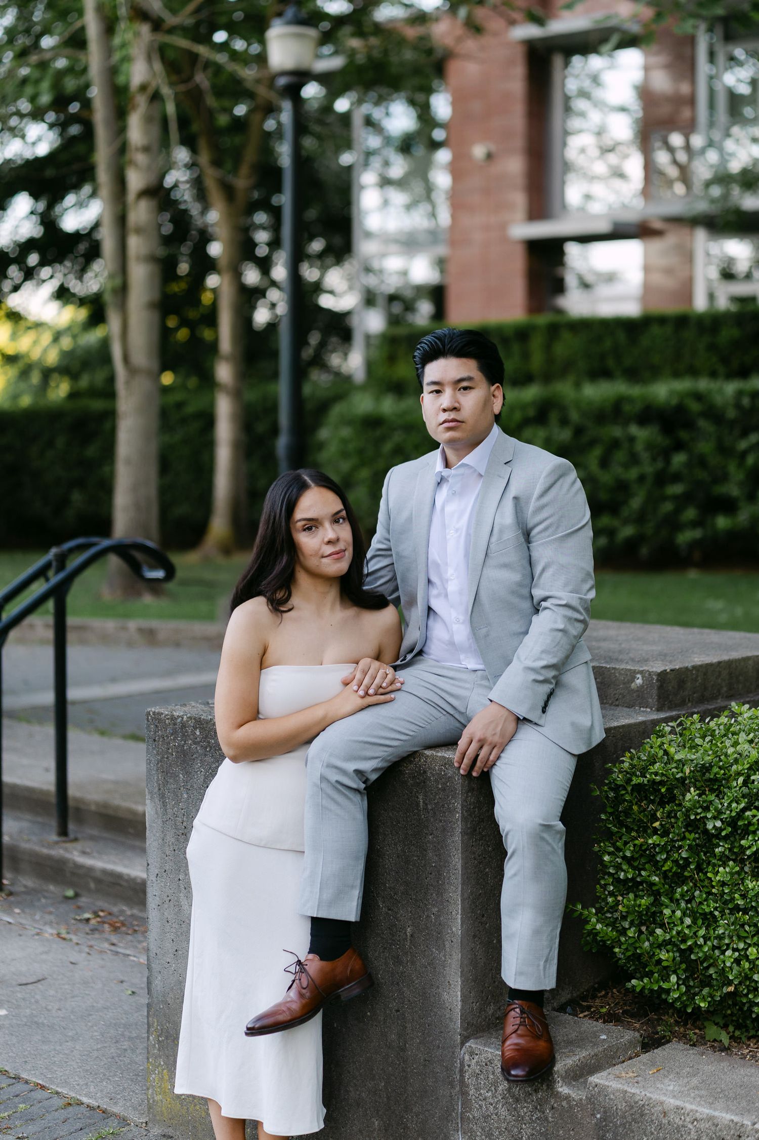 Engagement portrait of a couple posing on a stone ledge outdoors, she in a white dress and he in a gray suit with brown shoes.