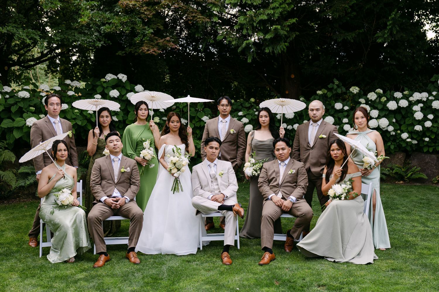 Wedding party poses in garden with white hydrangeas, bridesmaids in sage green gowns and groomsmen in brown suits.