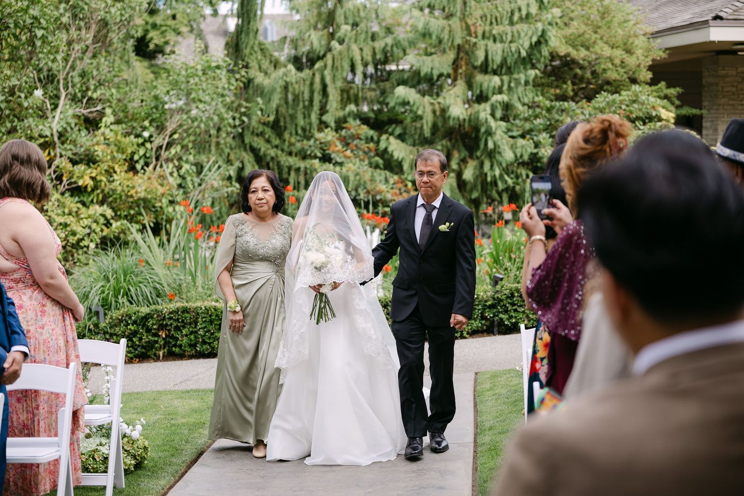 Bride walks down outdoor aisle with parents at garden wedding ceremony, wearing white gown and veil, holding white bouquet.