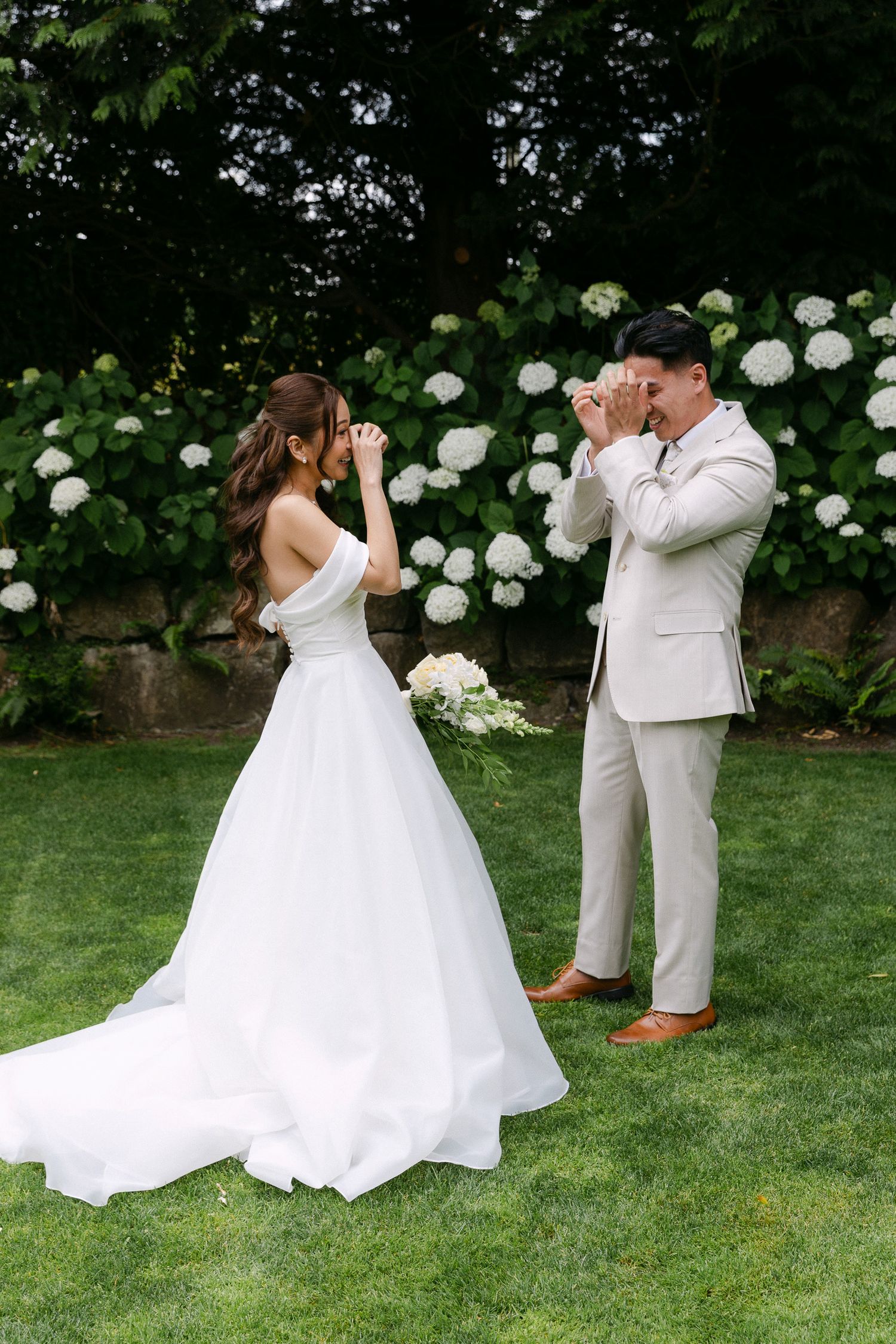 Groom in beige suit wipes tears seeing bride in white off-shoulder gown during first look in garden.