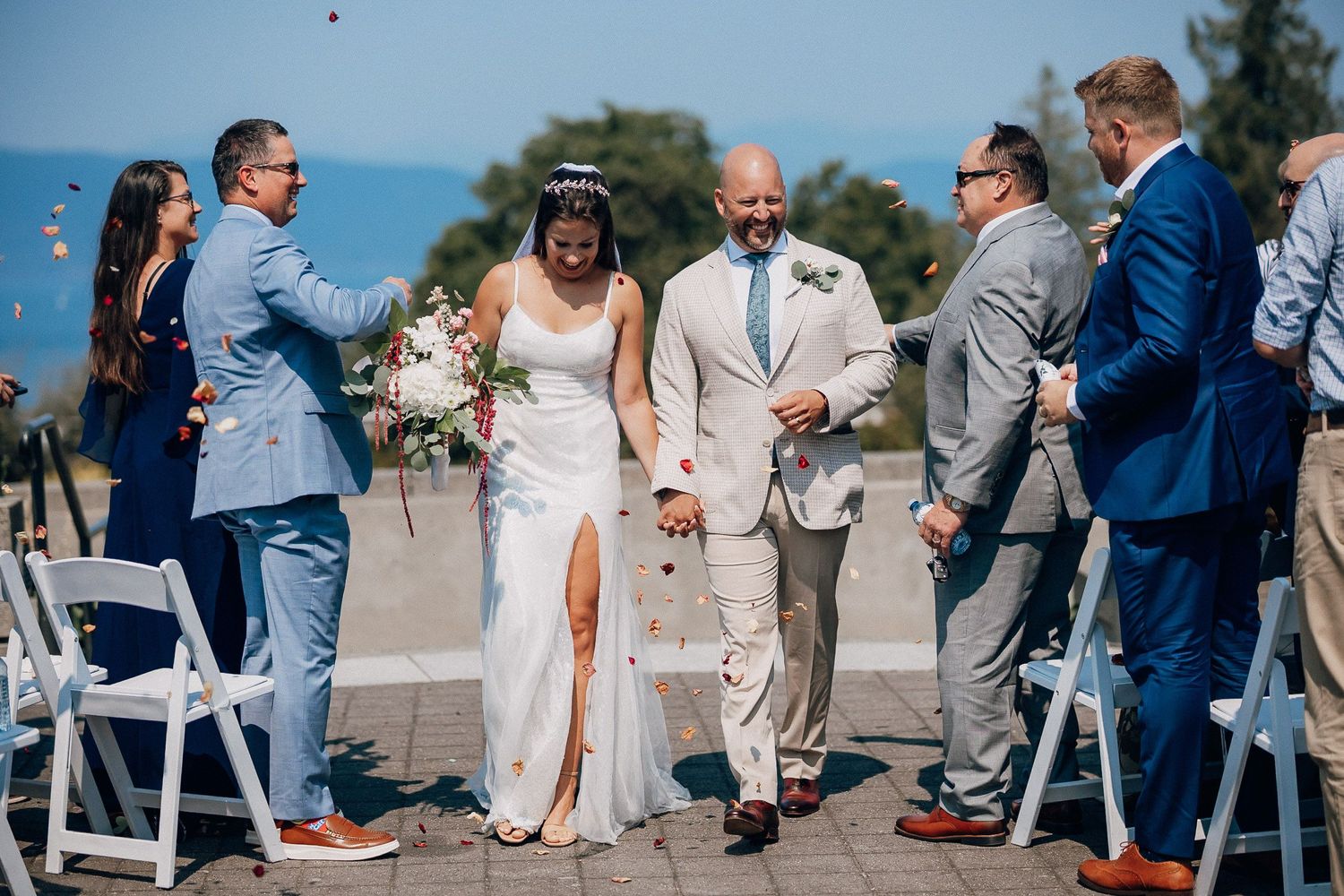 Wedding guests sit in white chairs on a beach during an outdoor seaside ceremony.