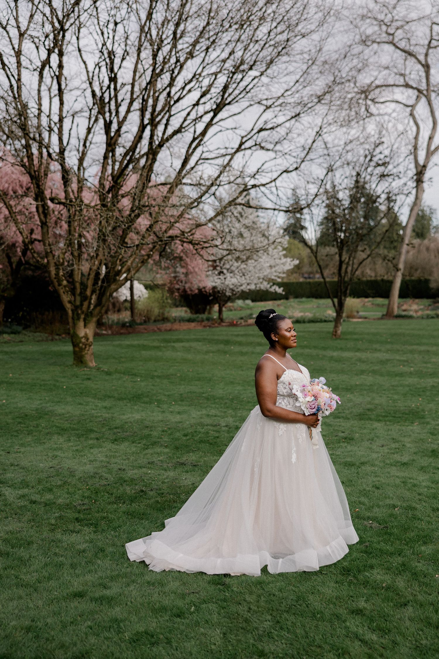 A bride in a flowing white dress poses in a spring garden with bare trees and green grass.