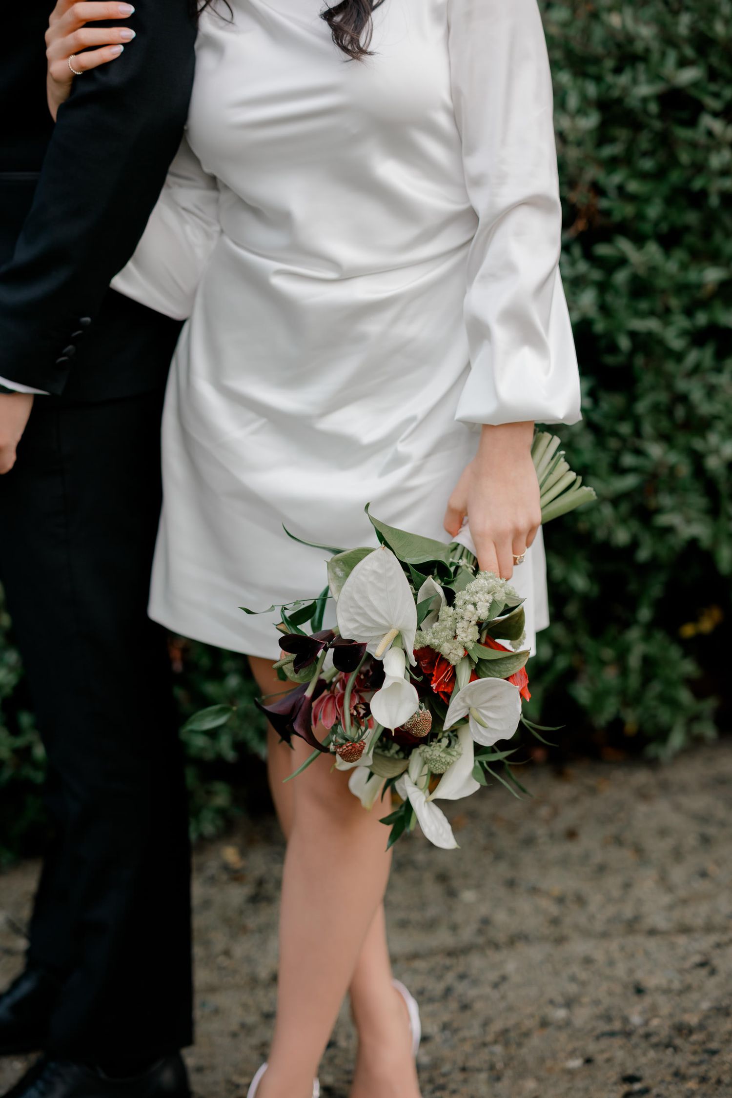 A person in a white dress holds a delicate bouquet of flowers with eucalyptus leaves against a garden background.