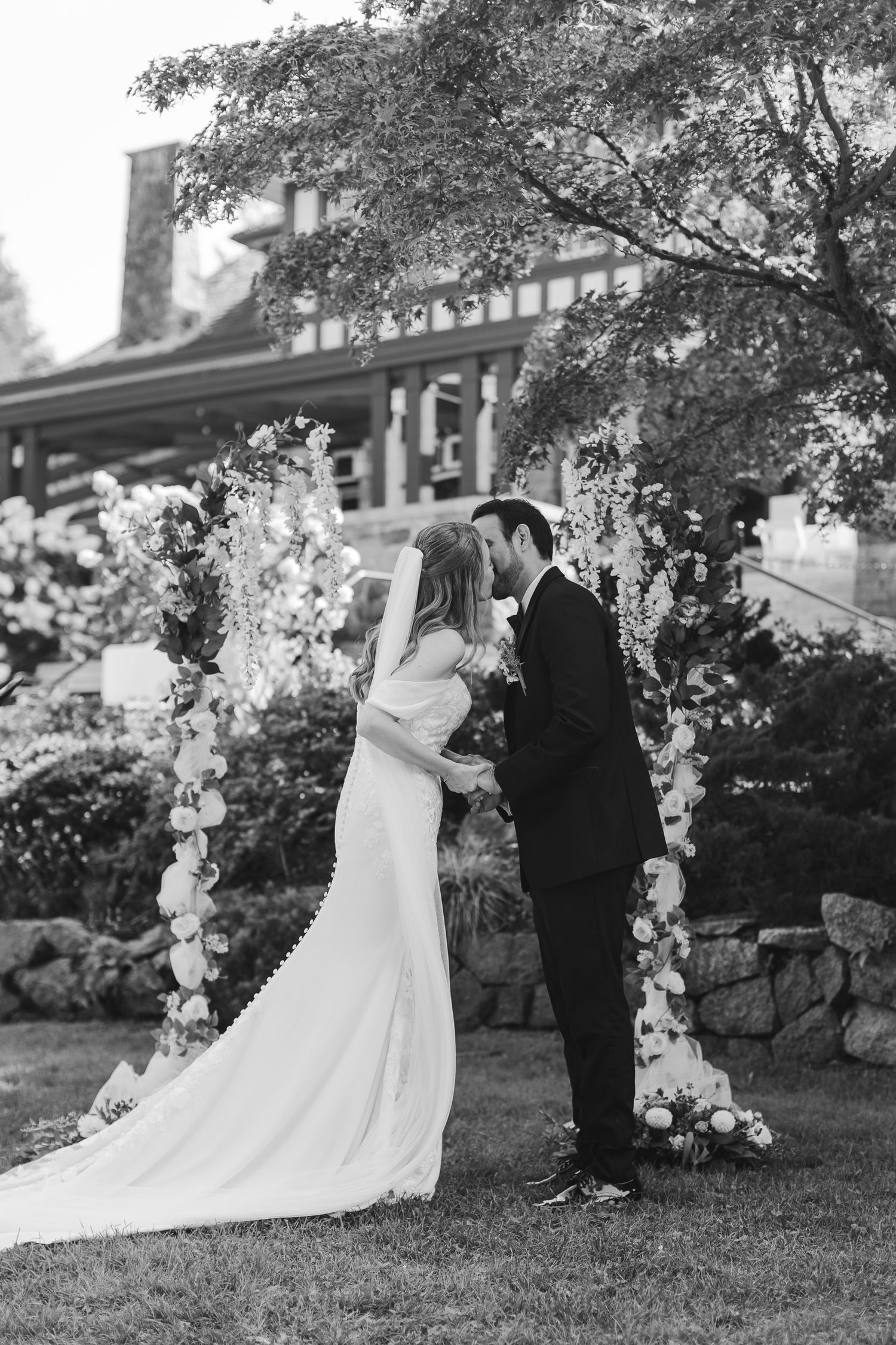 A series of black and white wedding photos showing a couple in formal attire under floral arches in an outdoor garden setting.