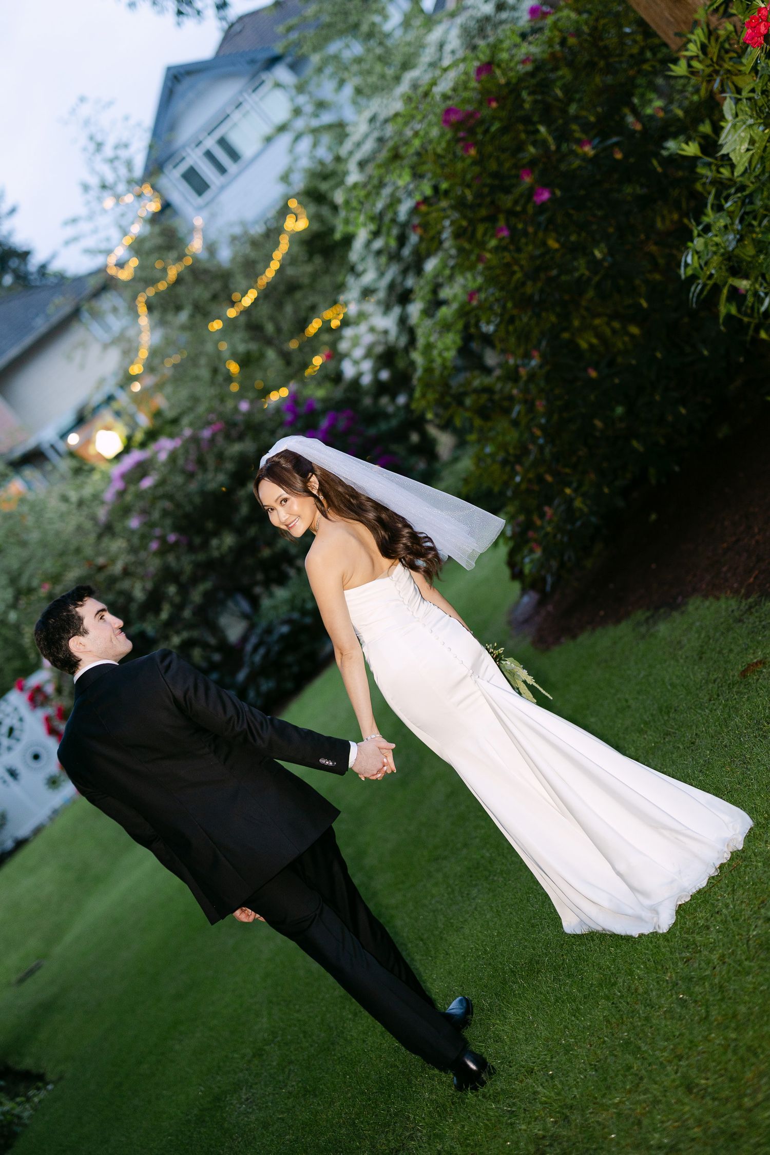 Bride and groom holding hands on green lawn at outdoor wedding, bride in white gown with veil, groom in black tuxedo.