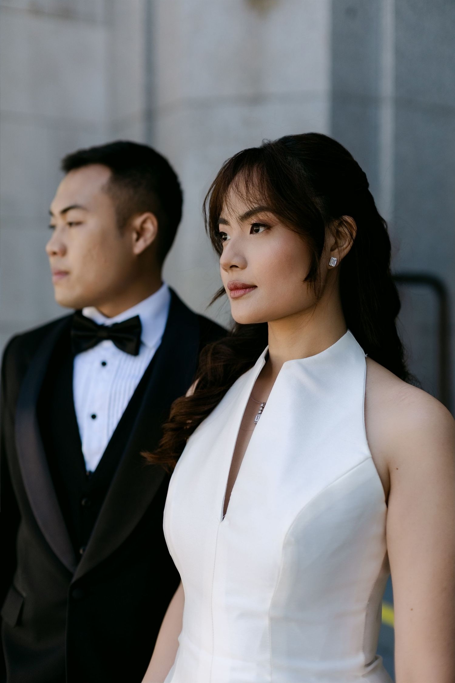 Elegant Asian couple in wedding attire, woman in white halter gown and man in black tuxedo, posing outdoors.