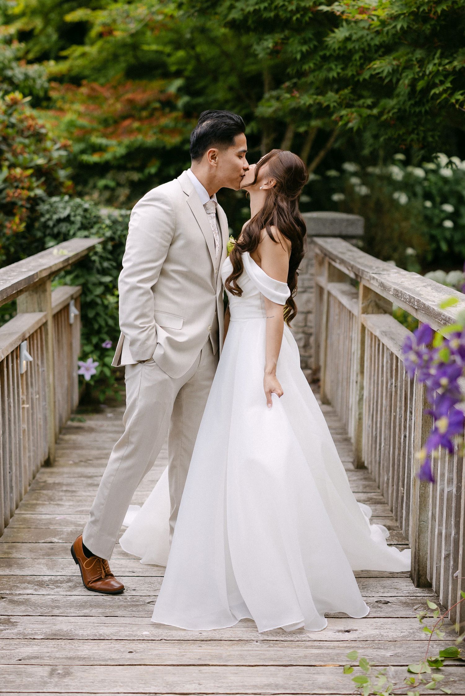 Bride and groom sharing a kiss on a wooden bridge surrounded by green trees, bride in off-shoulder white gown.