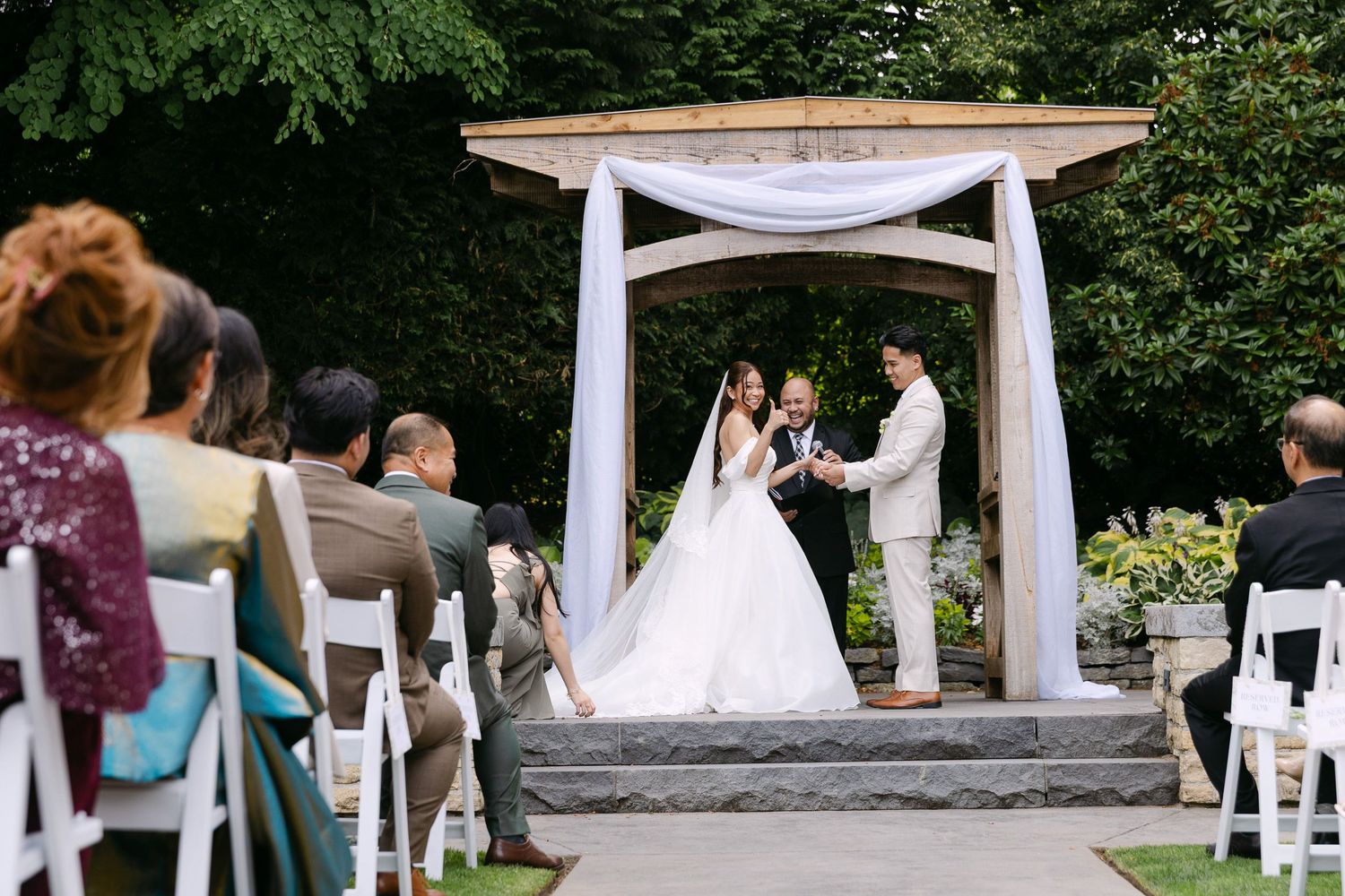 Outdoor wedding ceremony with bride and groom at floral arch, guests seated on white chairs in lush garden setting.