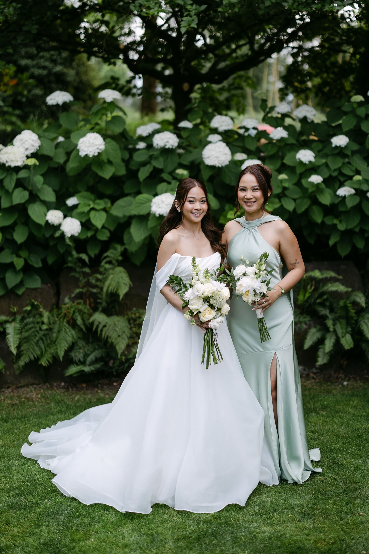Bride in off-shoulder white gown poses with bridesmaid in sage halter dress holding white bouquets outdoors.