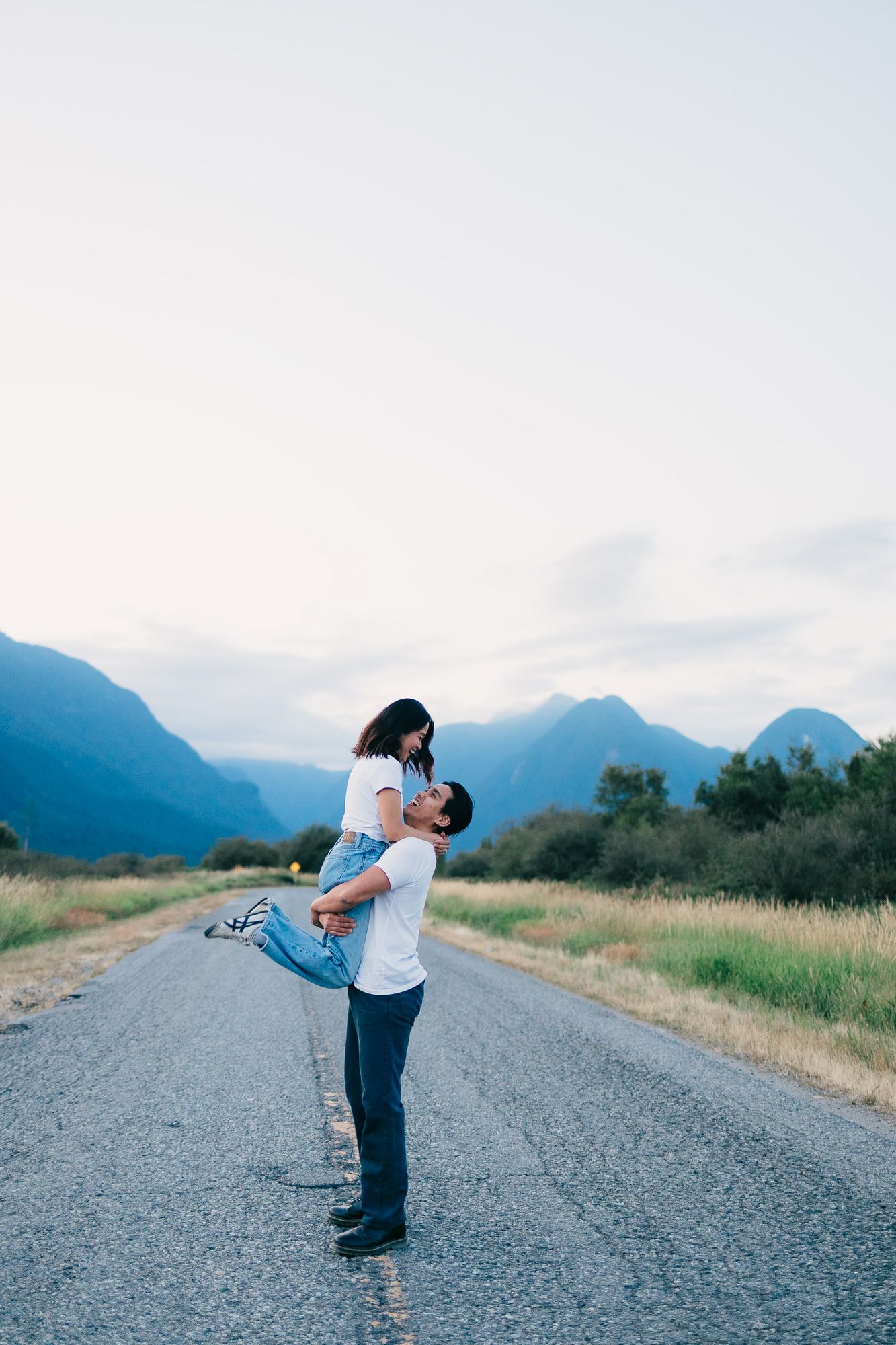 Couple embraces as a man lifts his partner off the ground on an empty road with mountain views at dusk.