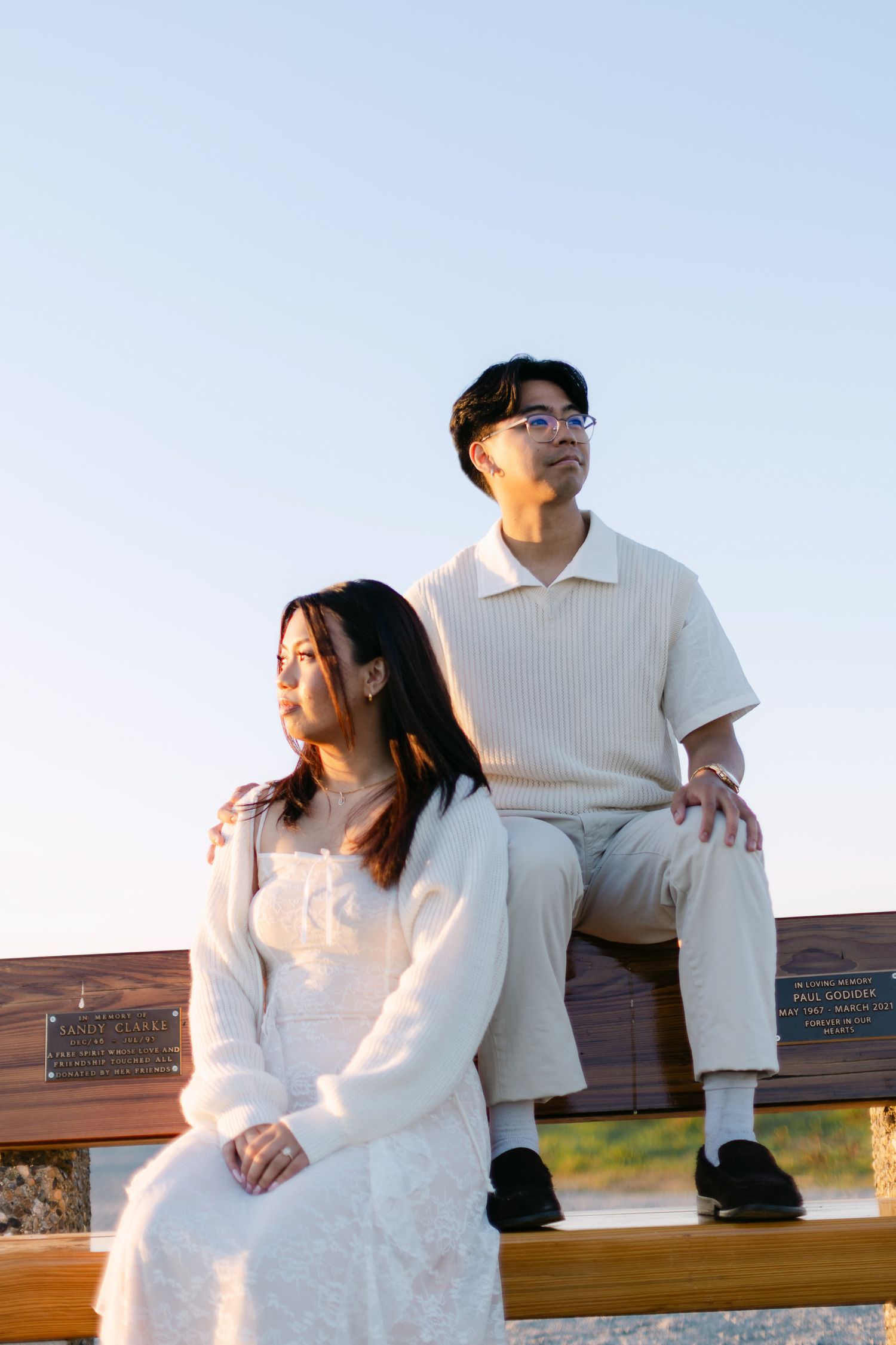 Couple in cream outfits sitting on a wooden memorial bench at golden hour, the man in glasses behind the woman in a white dress.