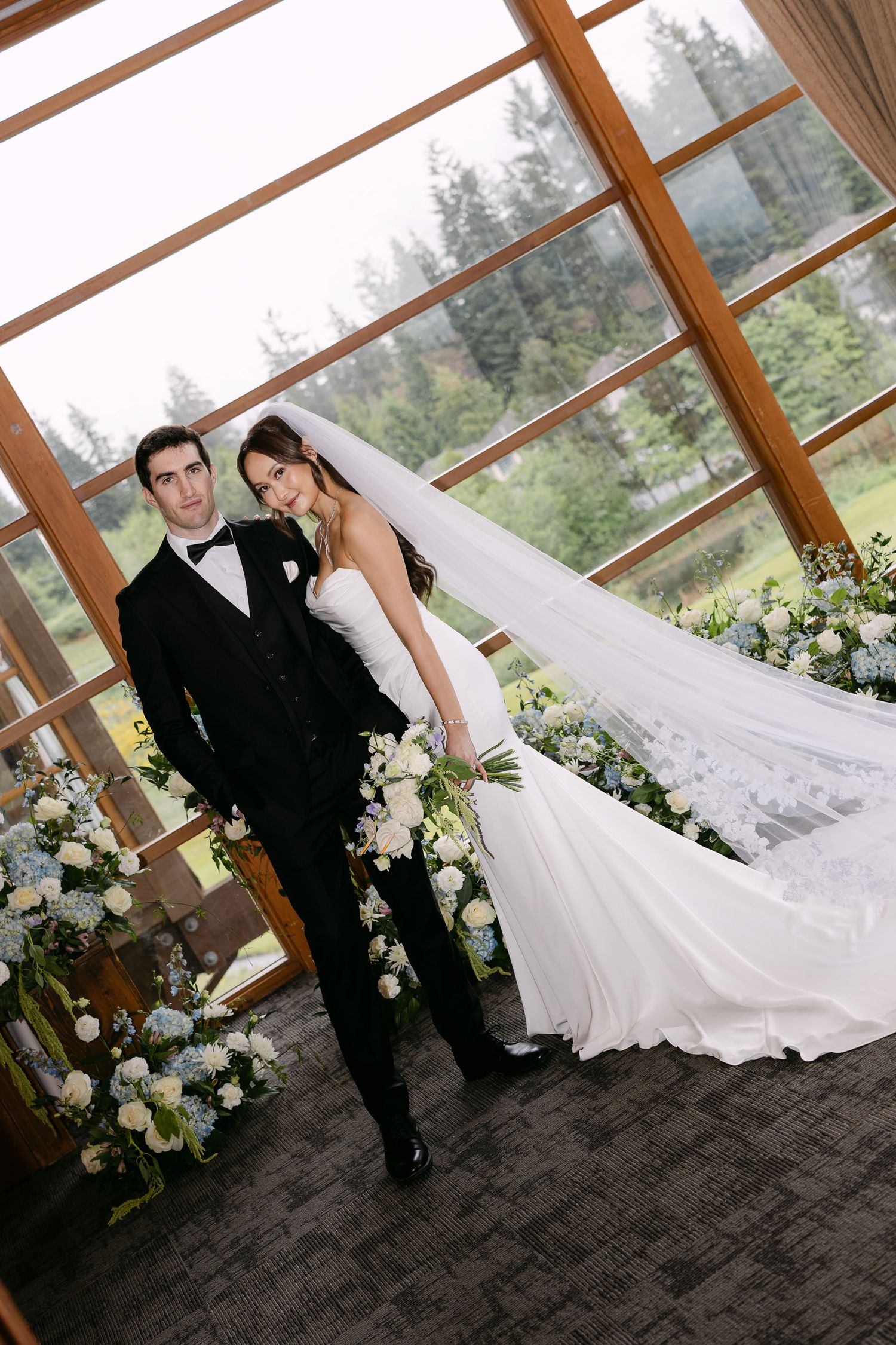 Bride in white gown with long veil and groom in black tuxedo pose together at elegant wedding venue with floral arrangements.