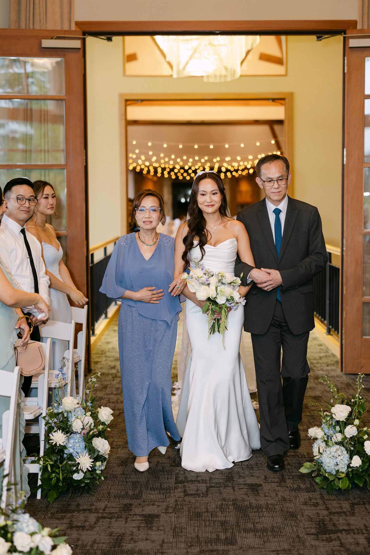 Bride walks down the aisle escorted by parents in elegant wedding ceremony with white floral arrangements.