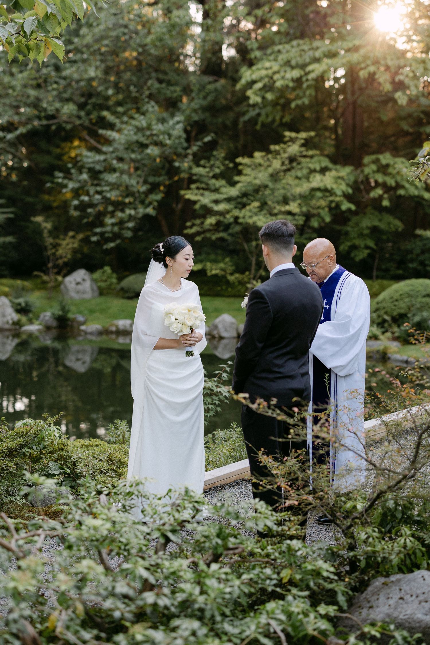Bride and groom exchange vows with officiant in a serene Japanese garden, surrounded by lush greenery and a tranquil pond.