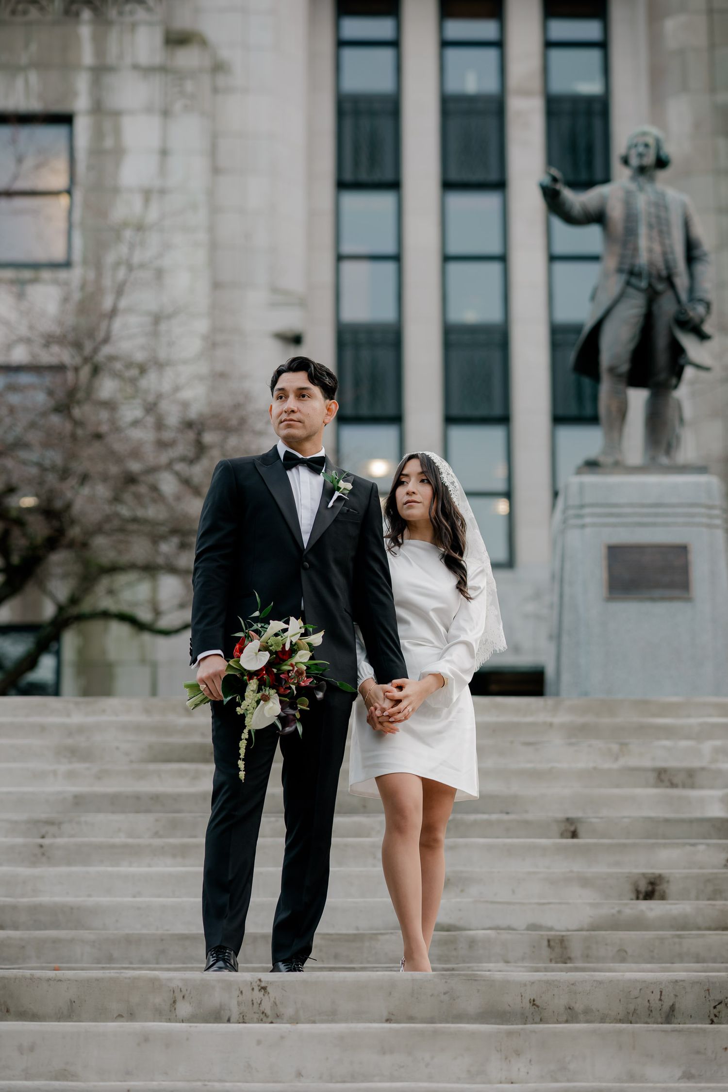Couple in wedding attire posing on grand stone steps of a historic building in black and white.