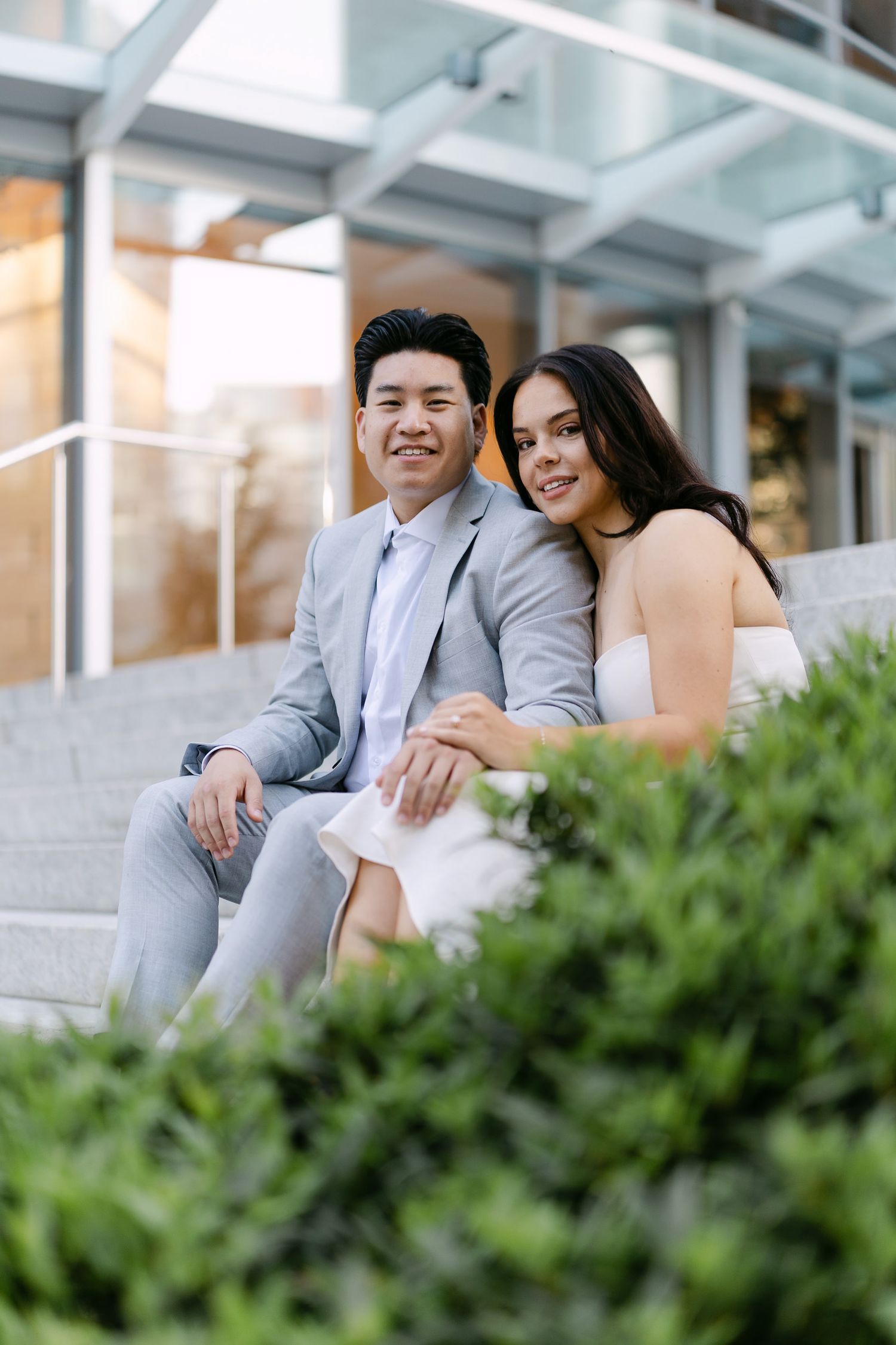 Smiling couple in light gray suit and white dress sit close together on outdoor steps behind green shrubs.