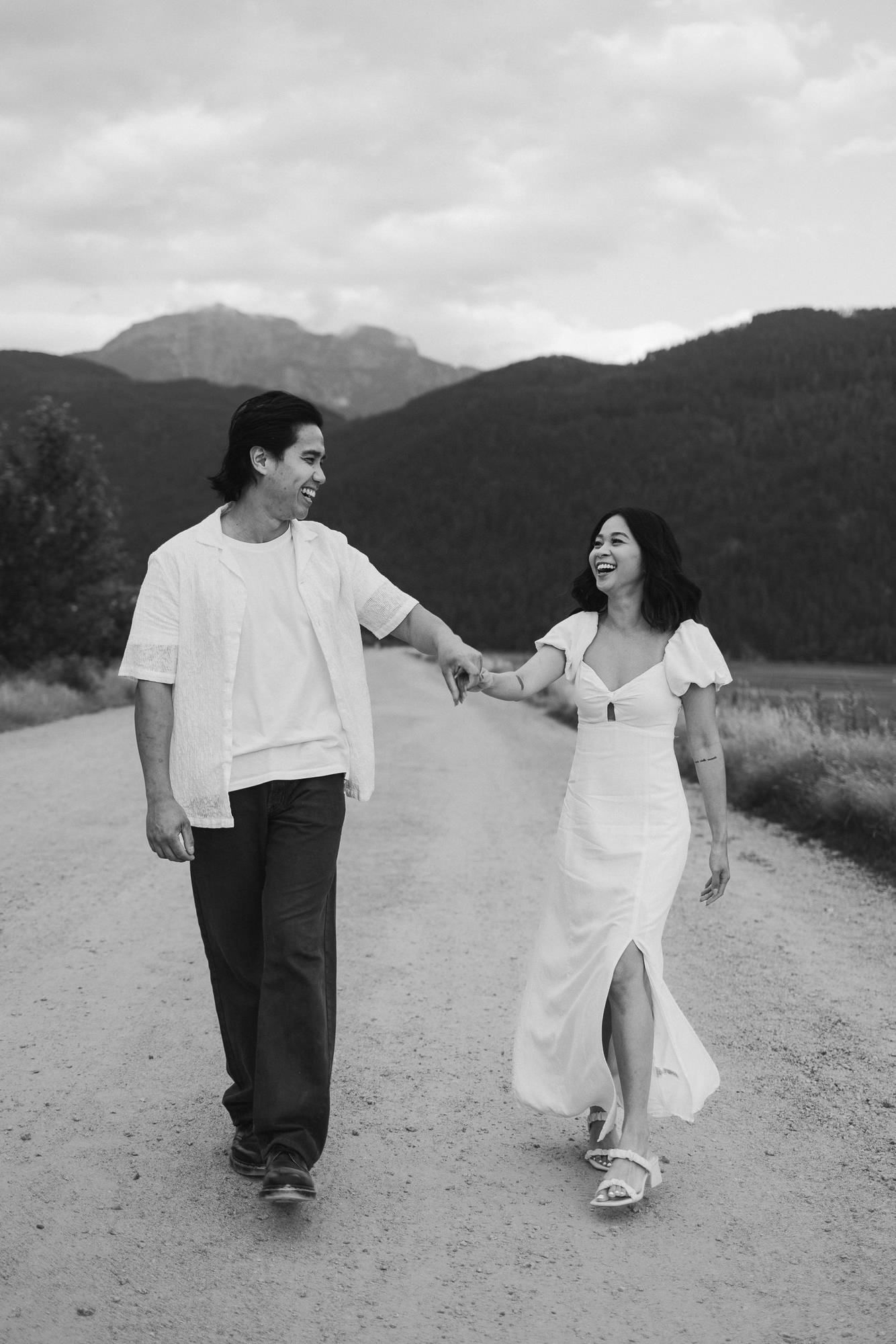 Black and white image of a joyful couple holding hands and walking down a dirt road with mountains in the distance.