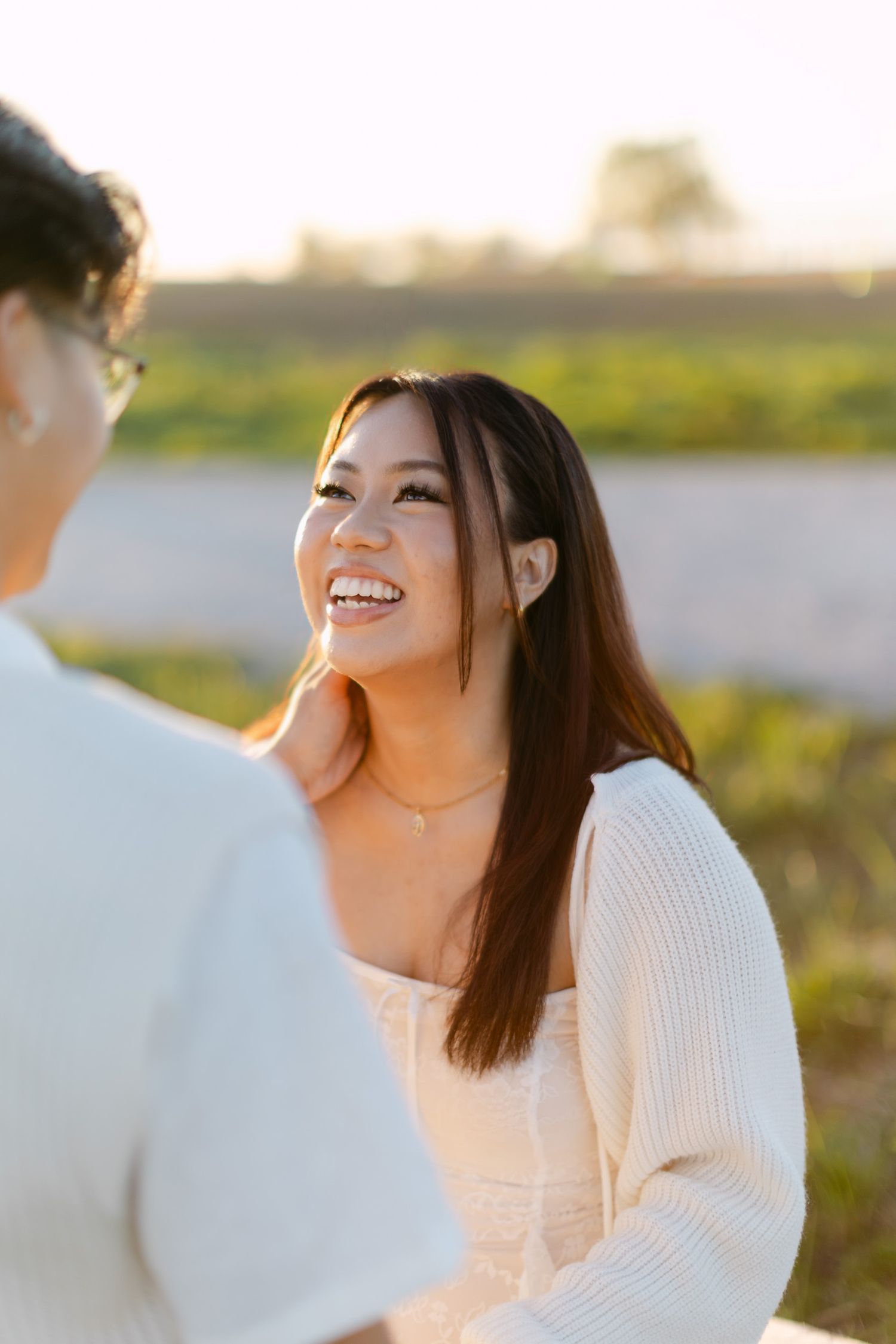 Laughing woman with long dark hair and a cream cardigan looks up at her partner in an outdoor golden hour portrait.