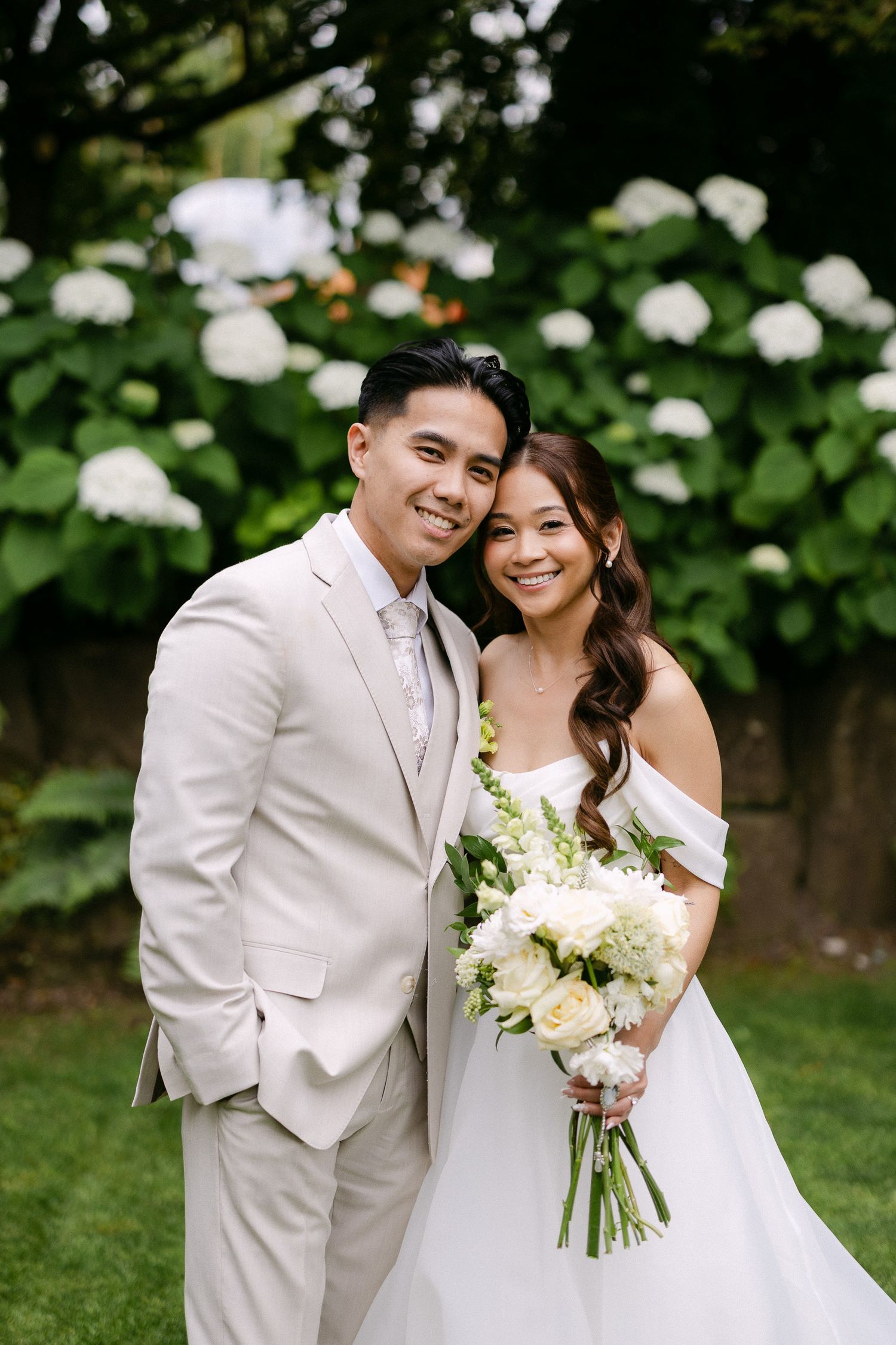 Smiling bride and groom pose together in garden, bride in white off-shoulder gown holding white bouquet.