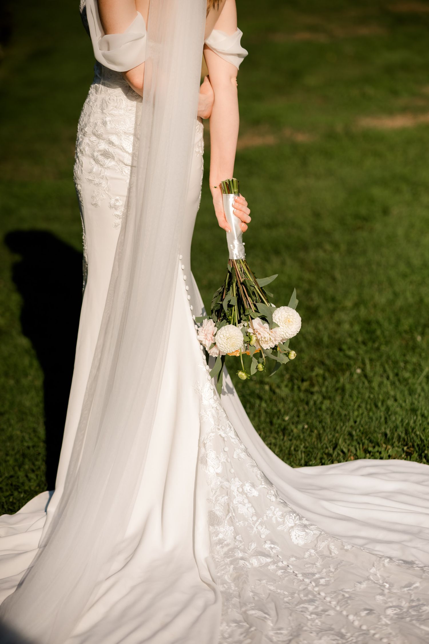 Elegant white lace wedding dress with flowing train and delicate bouquet of white flowers on green grass.
